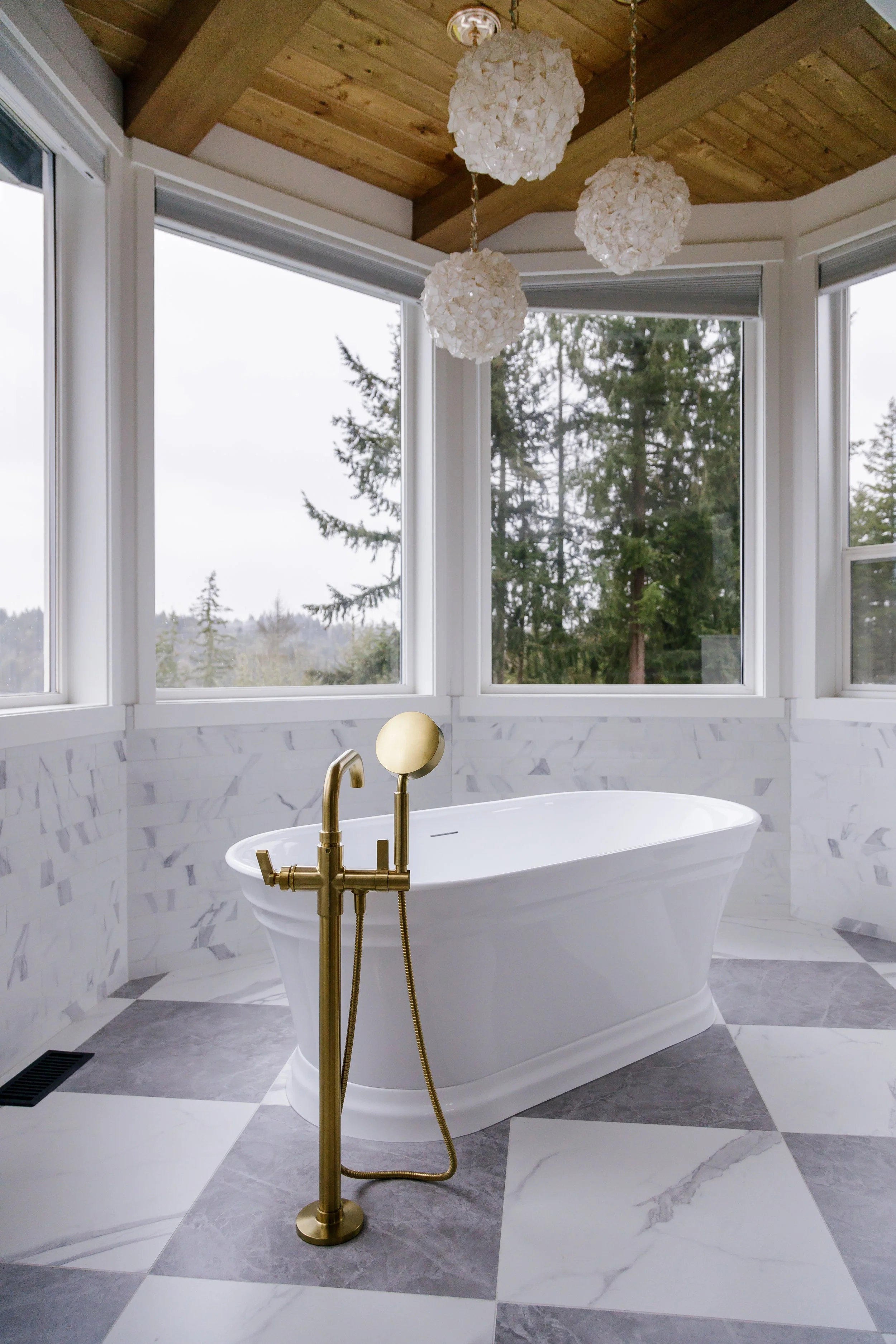 A modern bathroom with a freestanding bathtub, large windows showing trees outside, wooden ceiling, and hanging white pendant lights.
