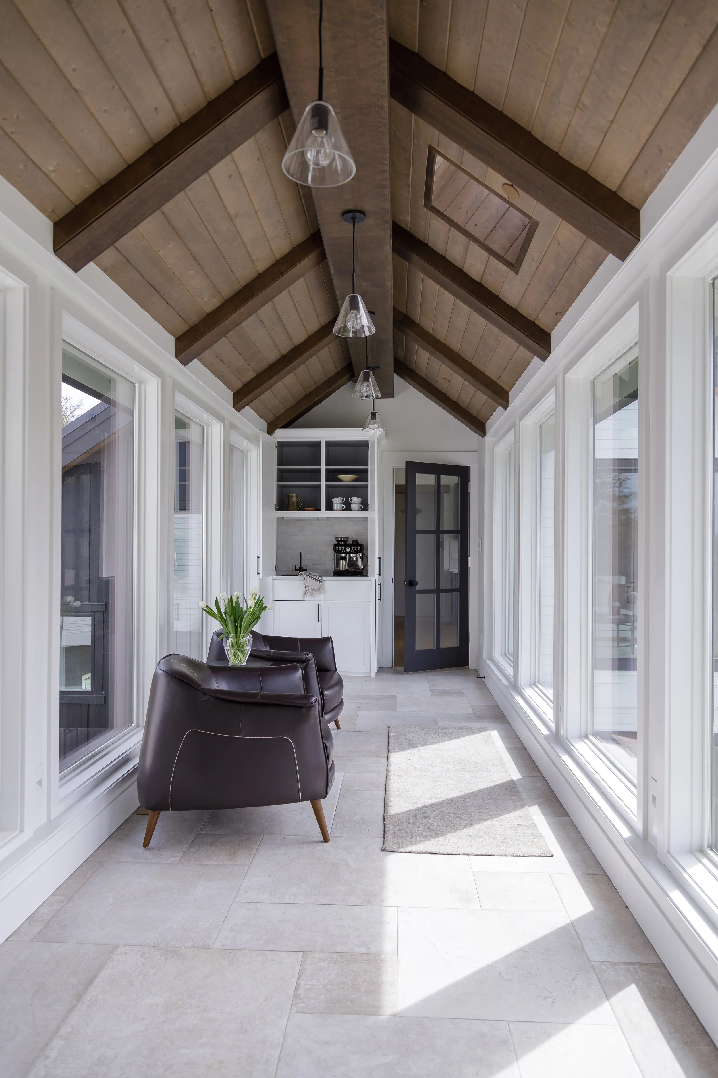 Sunlit enclosed porch or sunroom with large windows, wooden ceiling with exposed beams, two black leather armchairs, a small rug, a vase with tulips, and a kitchenette at the far end with open shelves and a coffee maker.