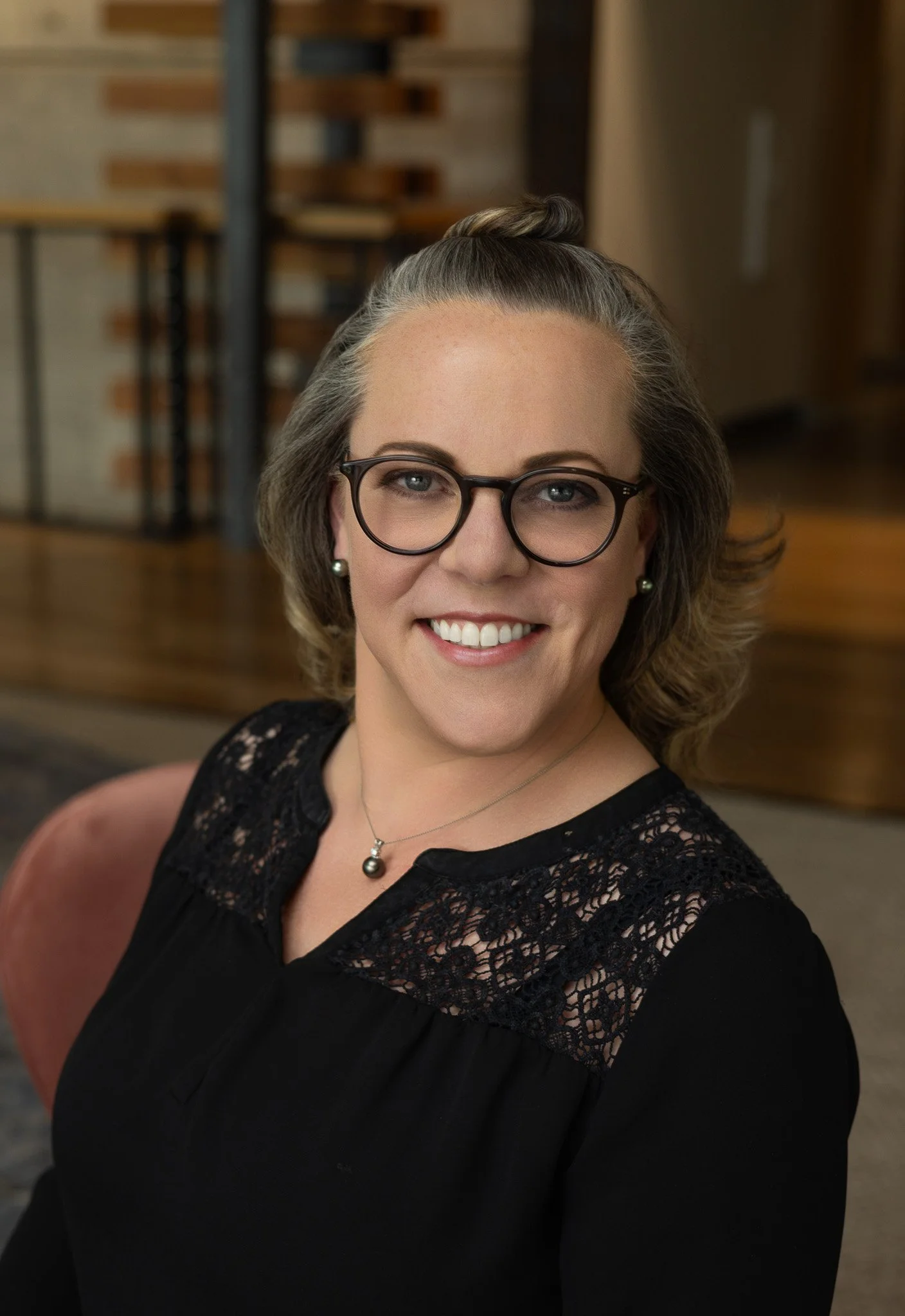 A woman with glasses, earrings, and a necklace smiling in an indoor setting.