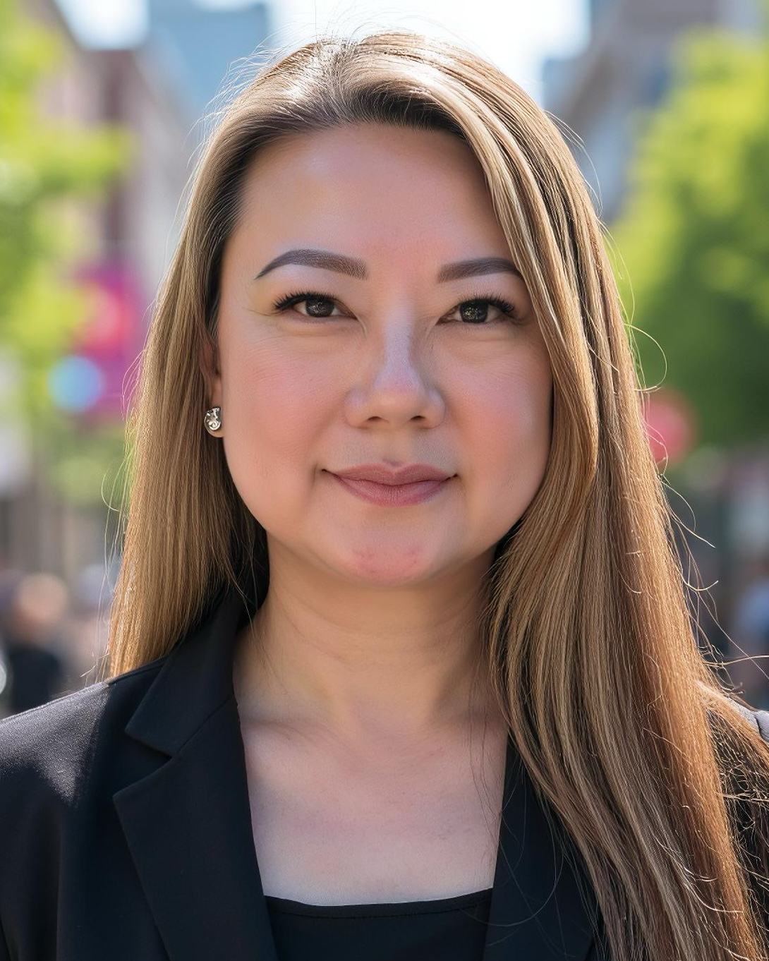 Close-up of a smiling woman with long light brown hair, wearing earrings and a black blazer, standing outdoors with trees and buildings in the background.
