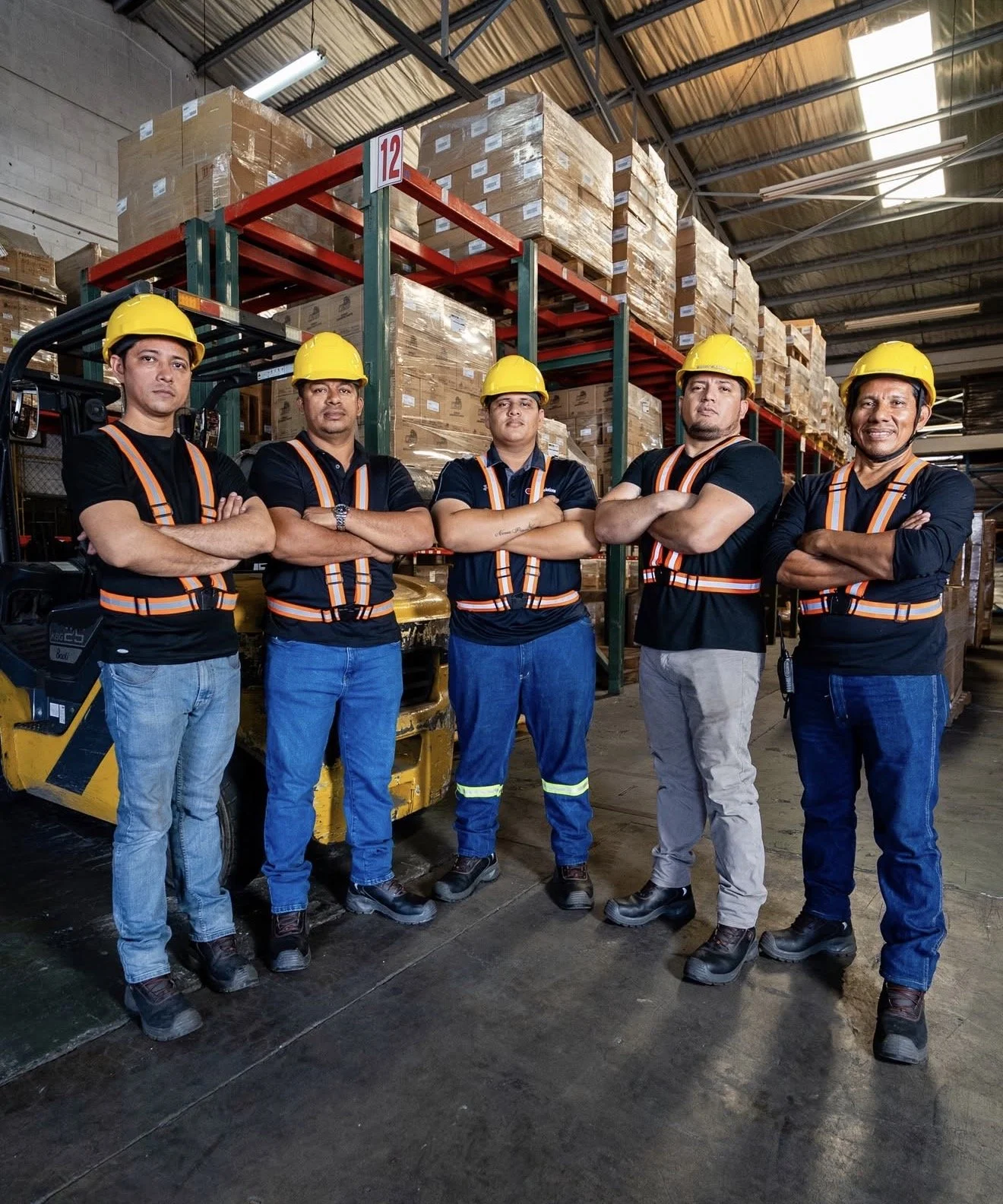 Five workers standing in a warehouse with arms crossed, wearing safety helmets, reflective vests, and work boots, with shelves of packed boxes behind them.