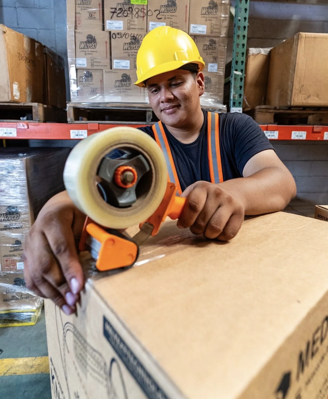 A worker in a warehouse wearing a yellow safety helmet and black t-shirt uses packing tape on a cardboard box.