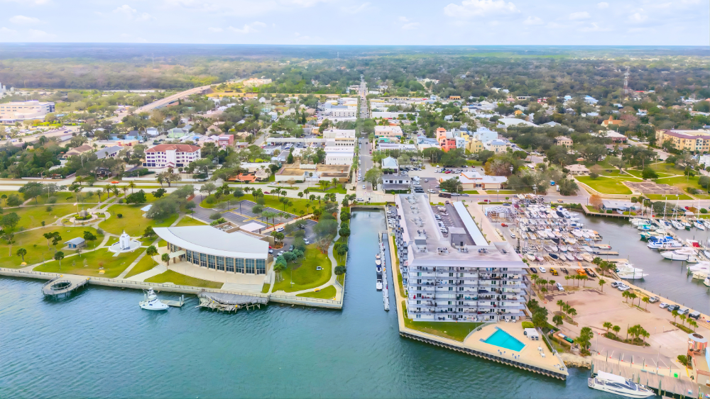 Aerial view of New Smyrna Beach, Florida, a coastal town with a marina, modern buildings, parks, and streets, along with water and boats docked at the harbor.