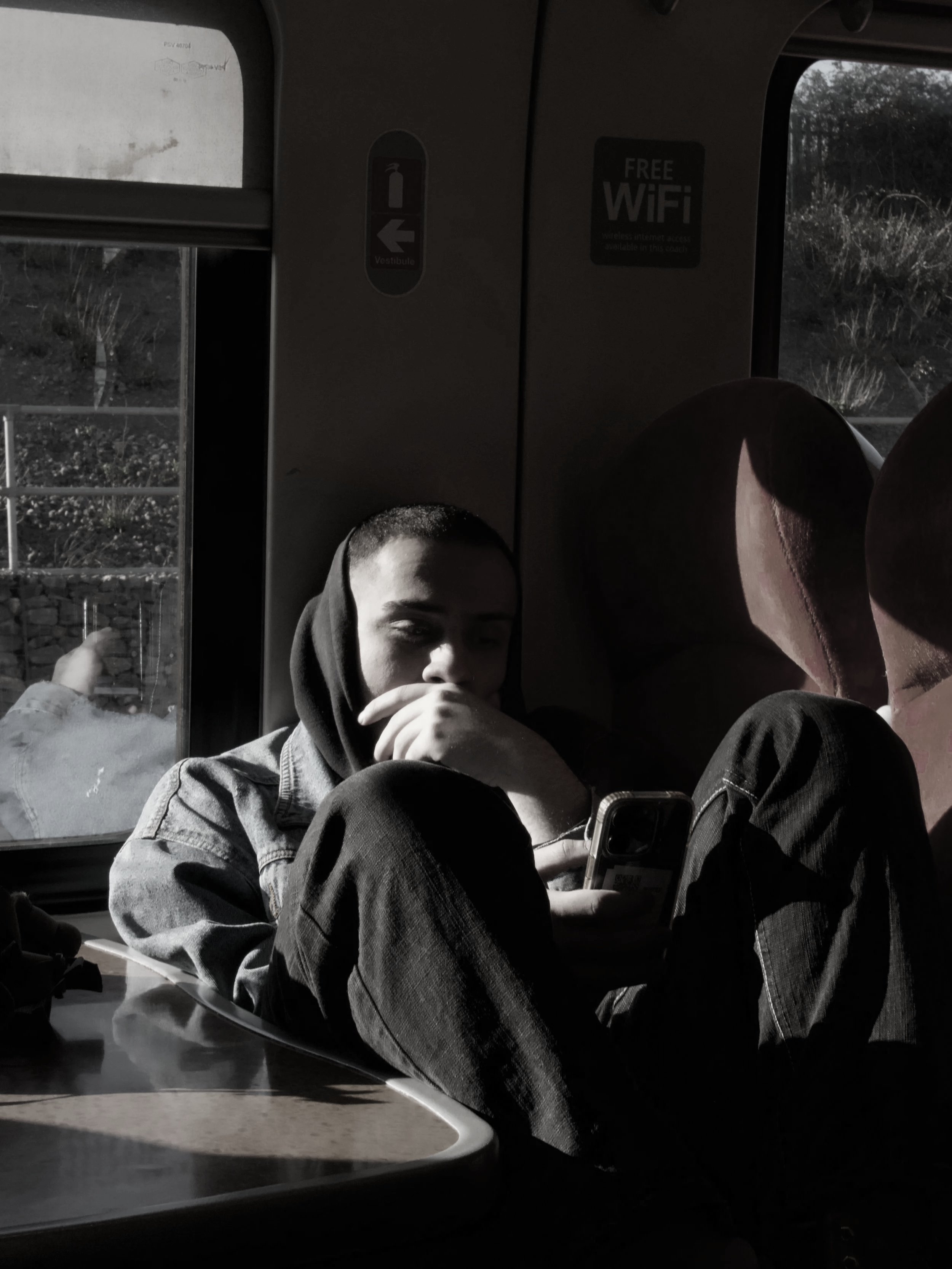 A young person with short hair, wearing a hoodie and denim jacket, sitting on a bus and using a smartphone, with sunlight streaming through the window.