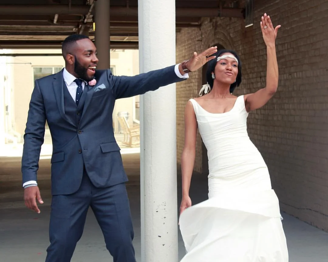 A joyful couple, the man in a navy suit and the woman in a white wedding dress, dancing and celebrating outdoors.