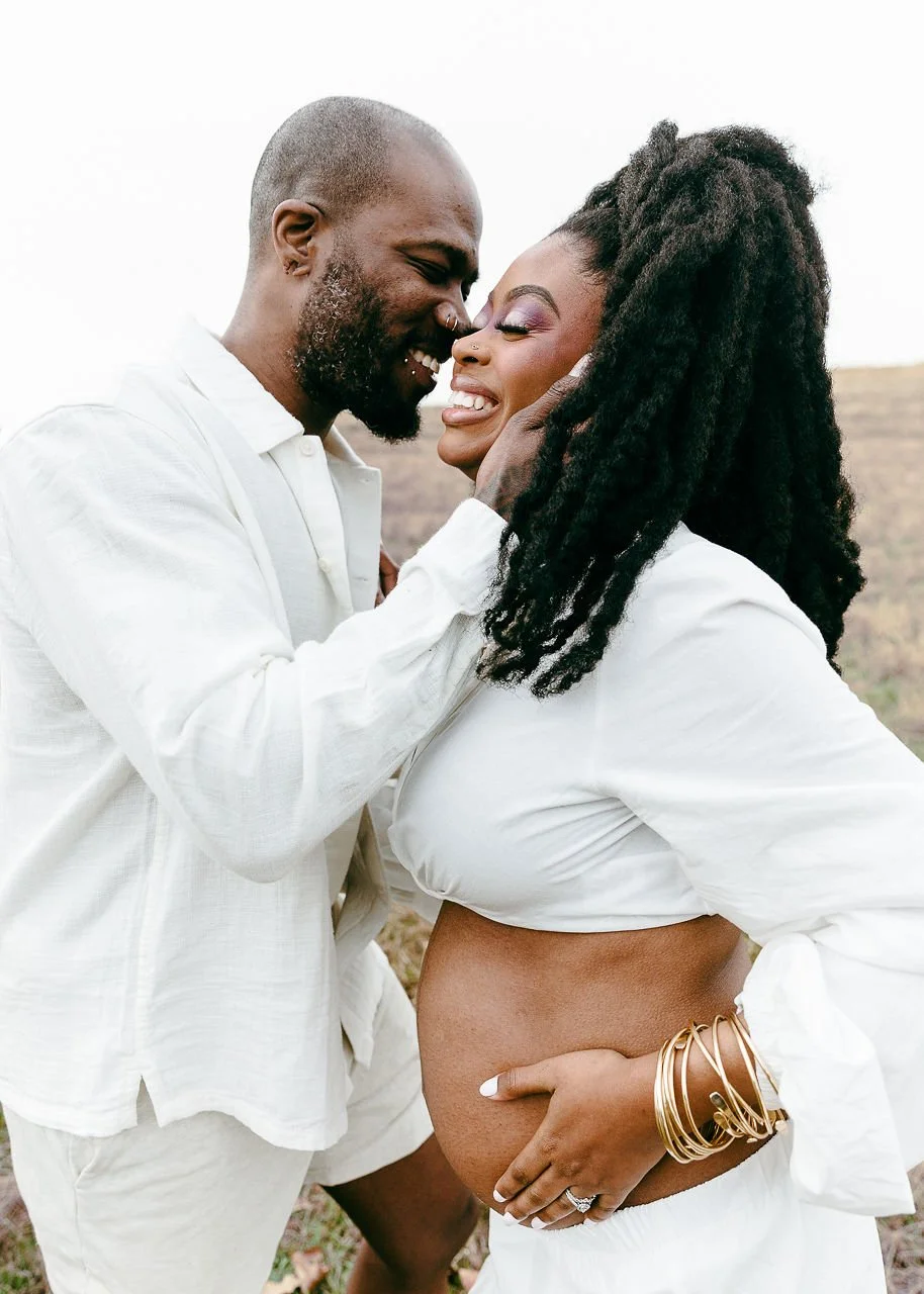 A couple smiling and touching foreheads outdoors, with the woman visibly pregnant, wearing white outfits and gold bracelets, both appearing joyful and intimate.