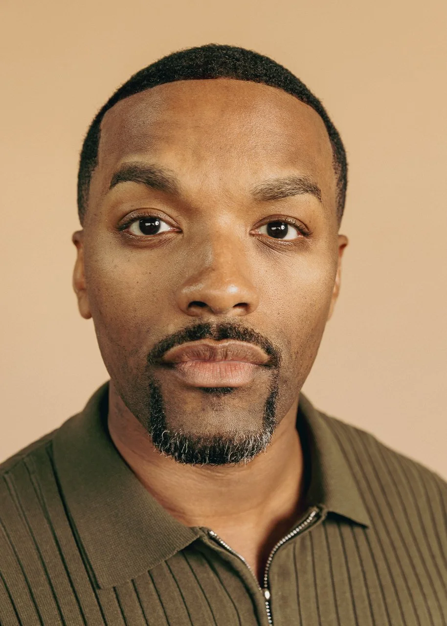Close-up portrait of a man with short curly black hair, a mustache and goatee, wearing a green striped shirt against a beige background.