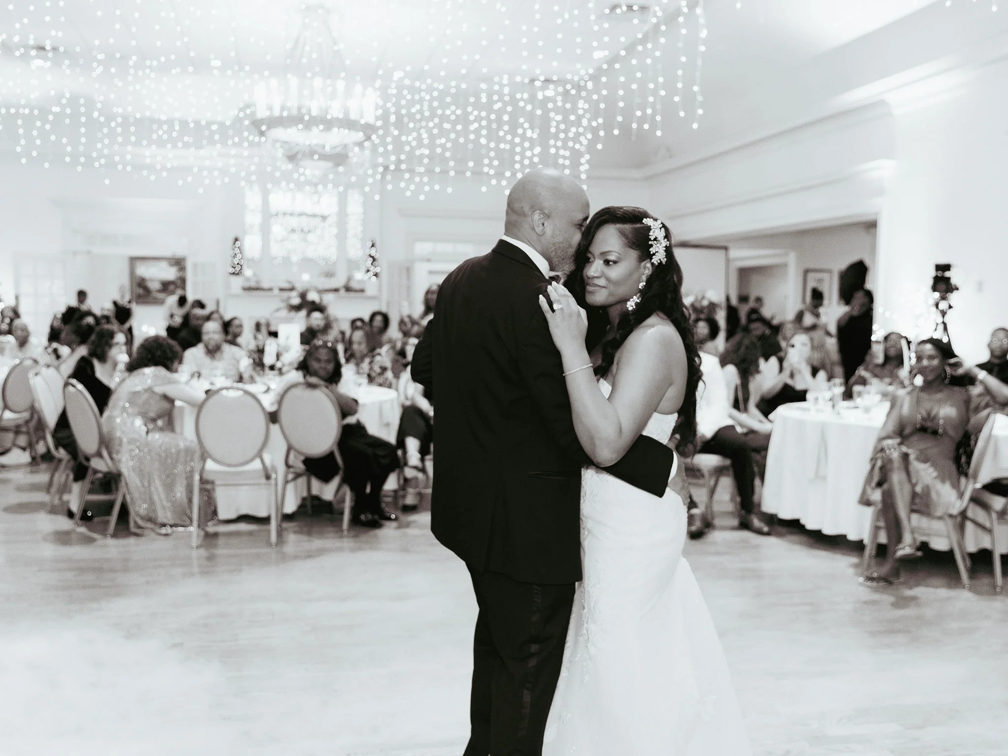 A bride and groom share their first dance at their wedding reception. The bride wears a strapless wedding gown with floral hair accessories, and the groom is in a black tuxedo. Guests are seated at round tables in the background under ceiling lights.