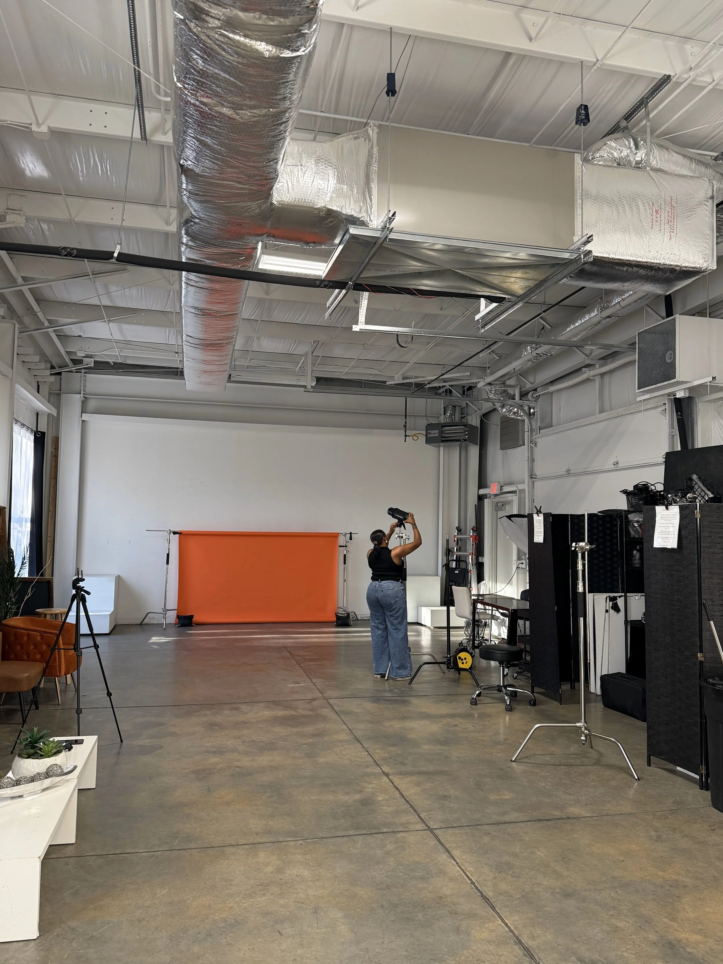 A woman standing in a photography studio with a large orange backdrop, holding a hairdryer, surrounded by lighting and photography equipment.