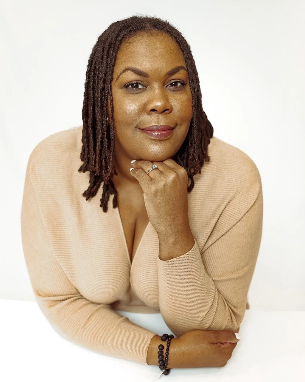 A woman with shoulder-length dreadlocks resting her chin on her hand, wearing a beige sweater, with a simple ring on her finger and a black beaded bracelet, smiling softly against a plain white background.