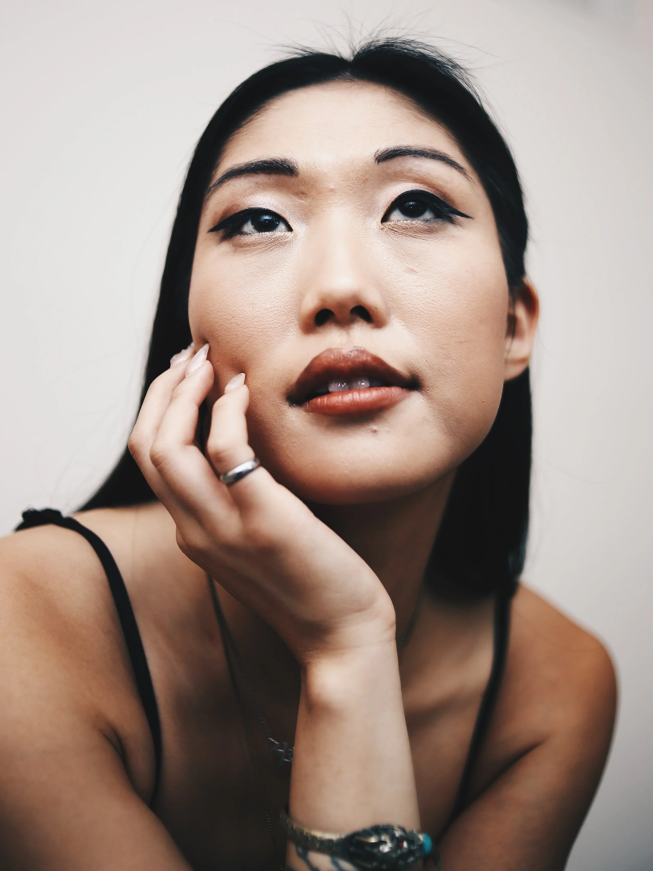 Close-up of an Asian woman with dark hair, wearing makeup, touching her face with her left hand, wearing a black strap top, a silver ring, a bracelet, and a necklace against a plain backdrop.