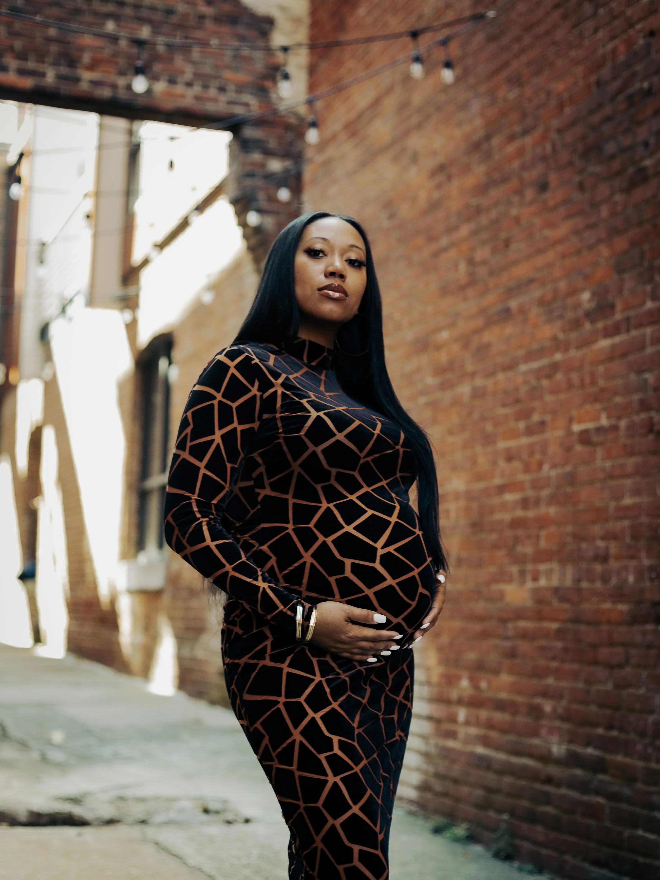 Pregnant woman standing in an alleyway with brick walls, wearing a black and brown geometric patterned dress, looking confidently at the camera.