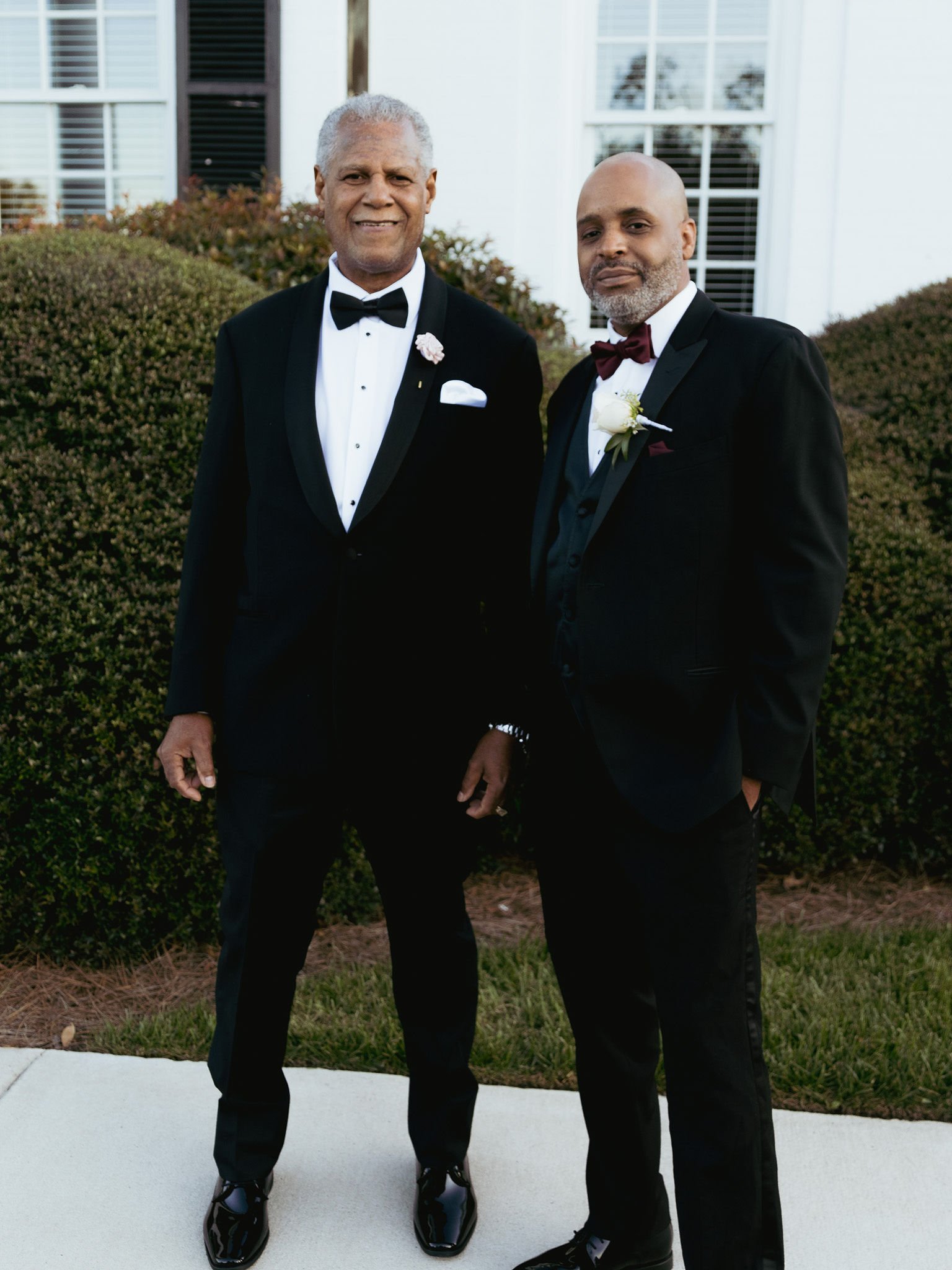 Two men dressed in black tuxedos and bow ties standing outdoors in front of bushes and a house, smiling at the camera.