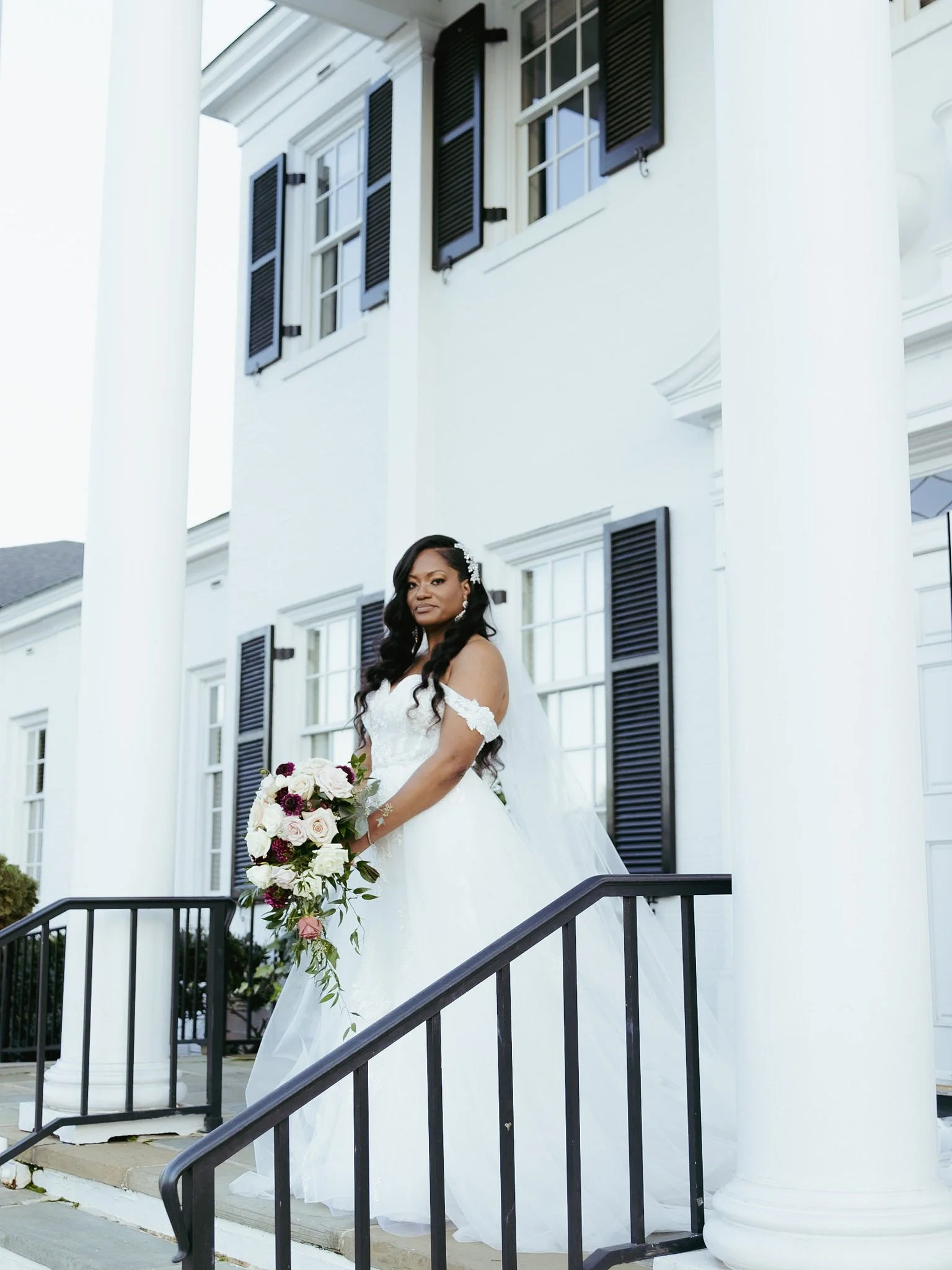 A bride in a white wedding gown holding a bouquet of flowers, standing on a front porch of a large white house with black shutters and columns.