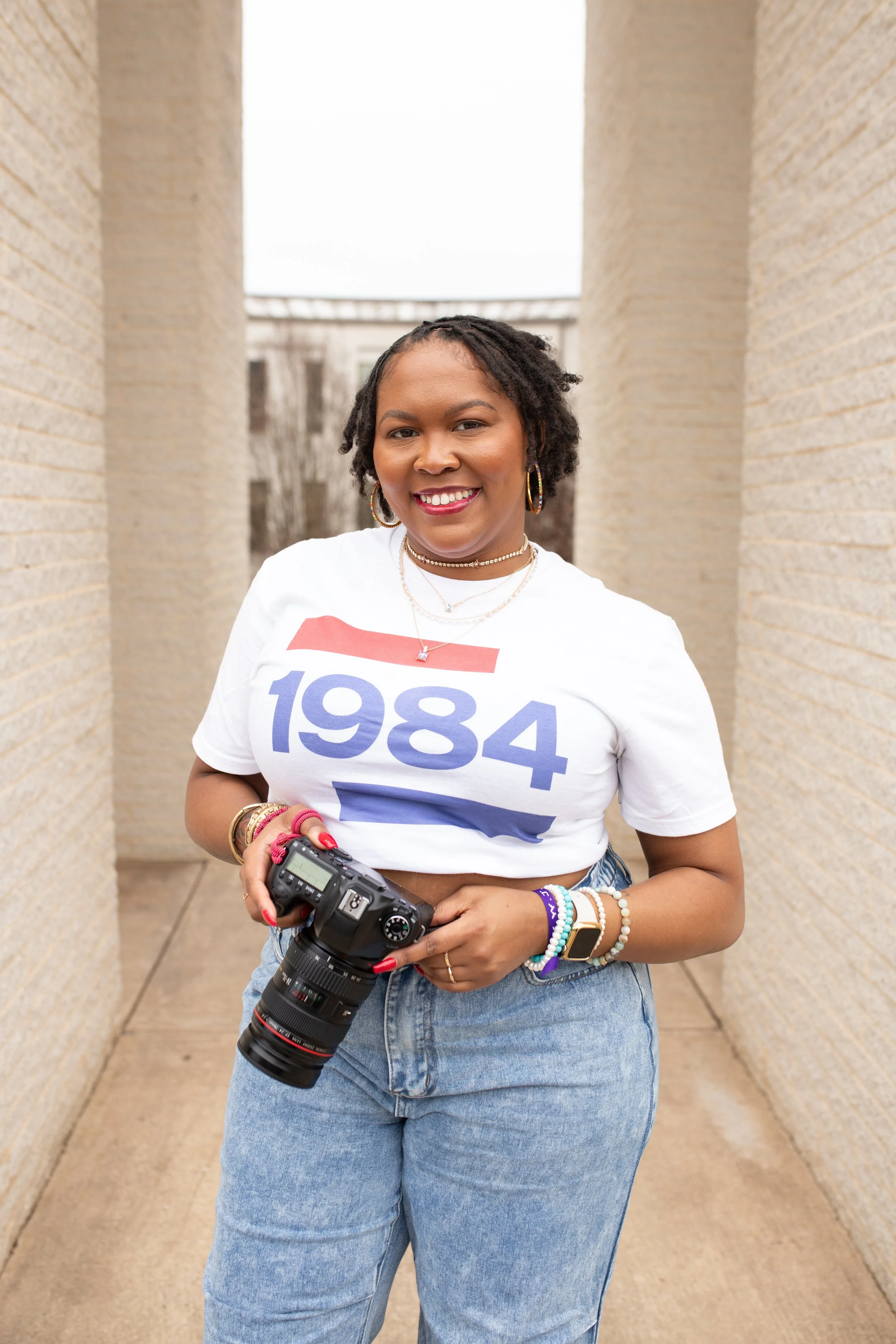 A woman with dark curly hair smiling, wearing a white T-shirt with '1984' printed on it, and holding a DSLR camera, standing between brick walls outdoors.