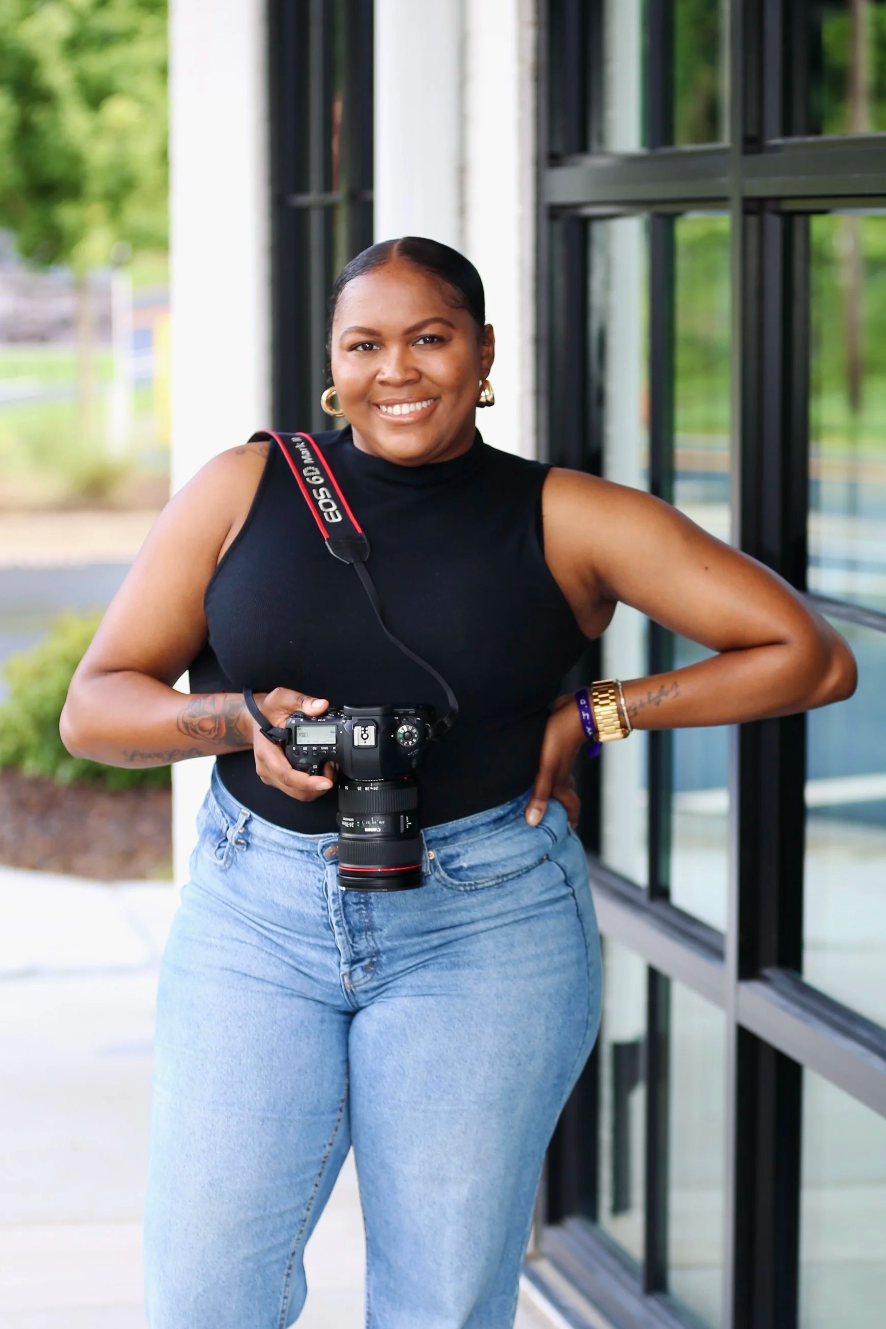 A woman standing outside near a building, smiling, holding a camera with a strap around her neck, wearing a black sleevelesstop, blue jeans, a gold watch, and hoop earrings.