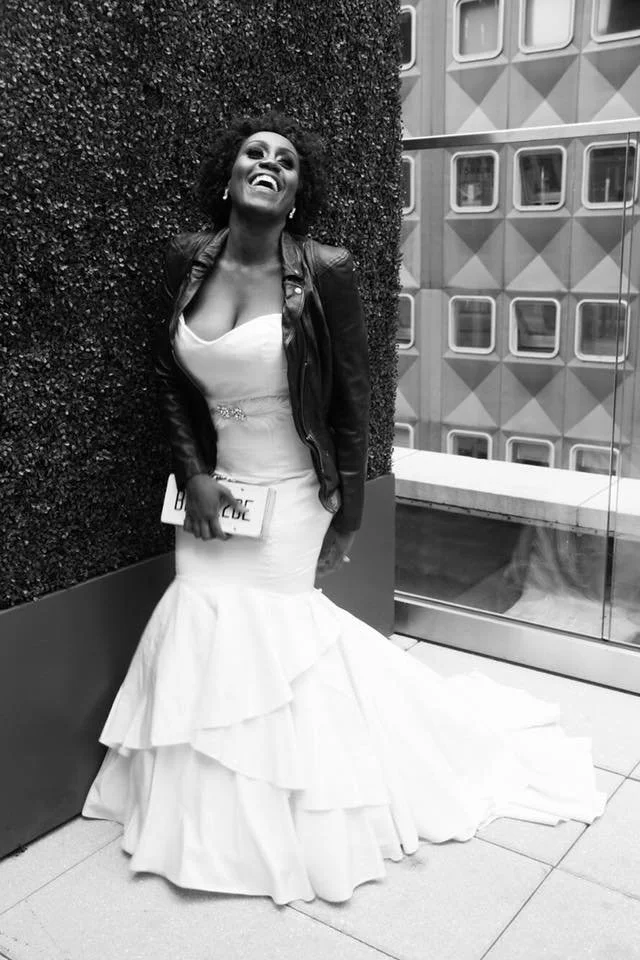 A woman in a white wedding dress and black leather jacket standing next to a textured wall, smiling and holding a clutch, outside a building with a glass entrance.