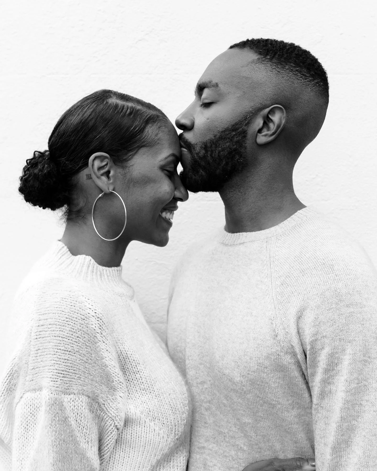 A black and white photo of a smiling woman with earrings and a bun, and a man with a beard, kissing their foreheads together against a plain wall.