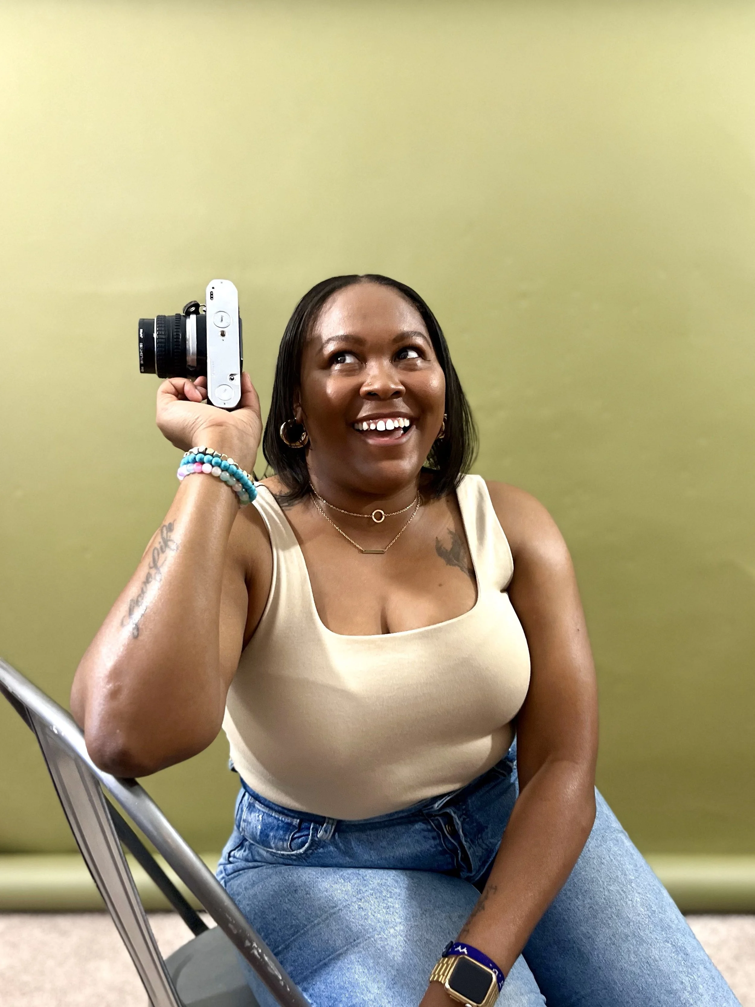 A woman with dark hair smiling, holding a camera to her head, wearing a beige tank top, blue jeans, and jewelry, sitting against a green wall.