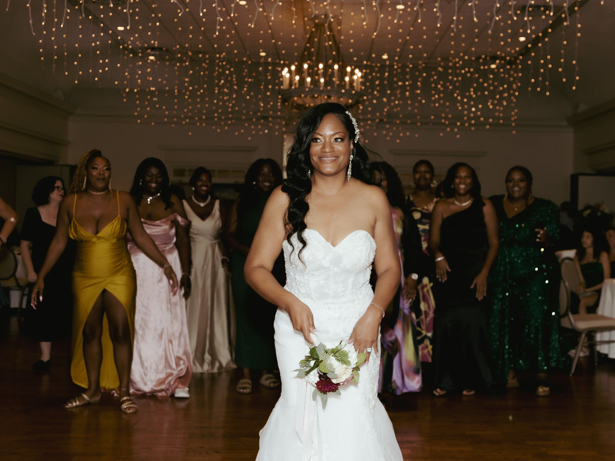 Bride in a white strapless wedding gown holding a bouquet, smiling, at a wedding reception with women in colorful dresses cheering in the background, decorated with string lights overhead.