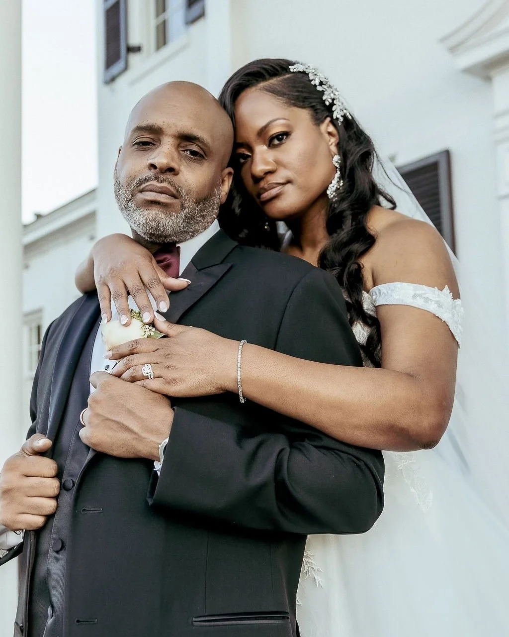 A bride and groom pose closely for a wedding photo outside a white building, with the bride resting her head on the groom's shoulder.