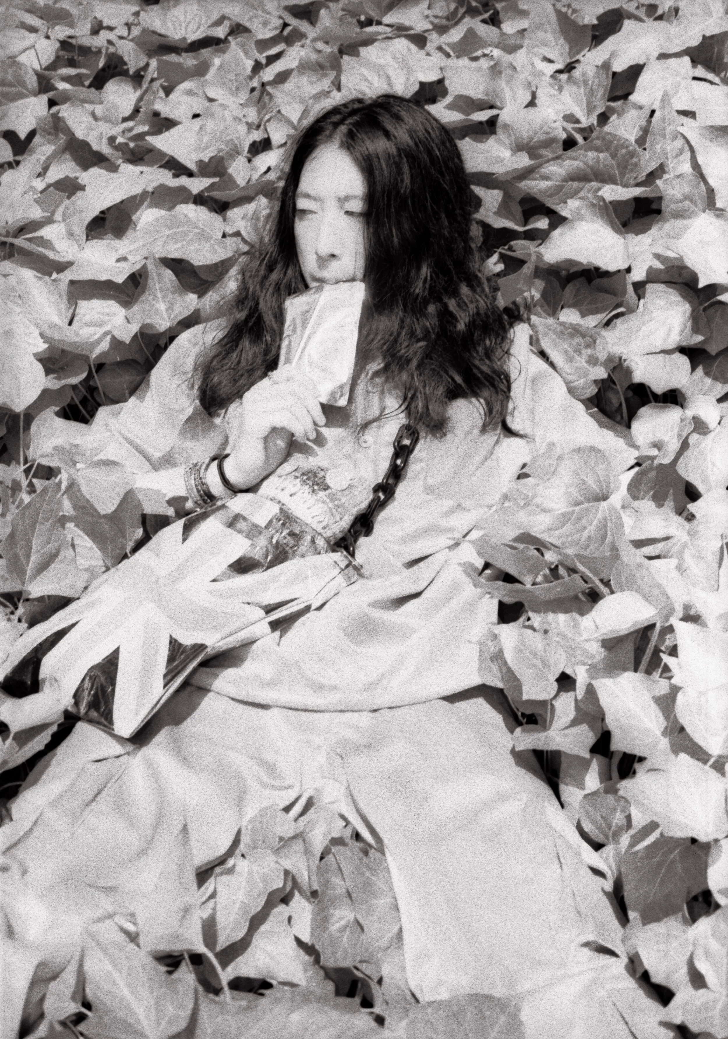 A woman with long wavy hair lying on a bed of large leaves, holding an ice pack near her mouth, wearing casual clothing and a bracelet.