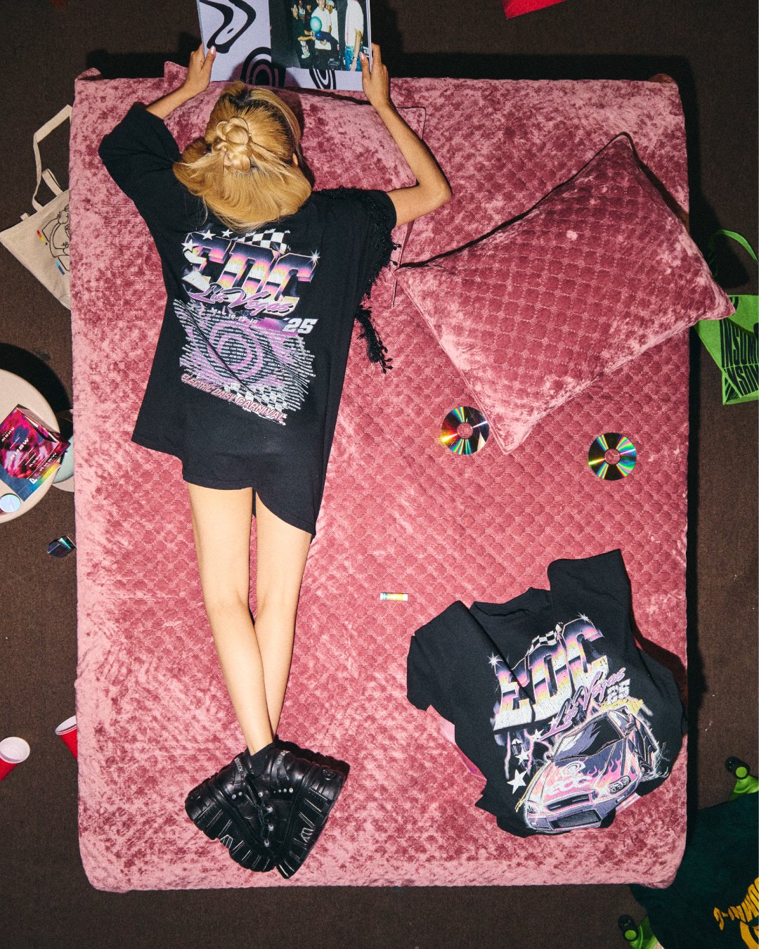 A person lying on a pink velvet bed, wearing a black oversized T-shirt with a retro design, black platform shoes, and blonde hair styled in a braid. The bed has matching pillows, with CDs and clothing scattered around.