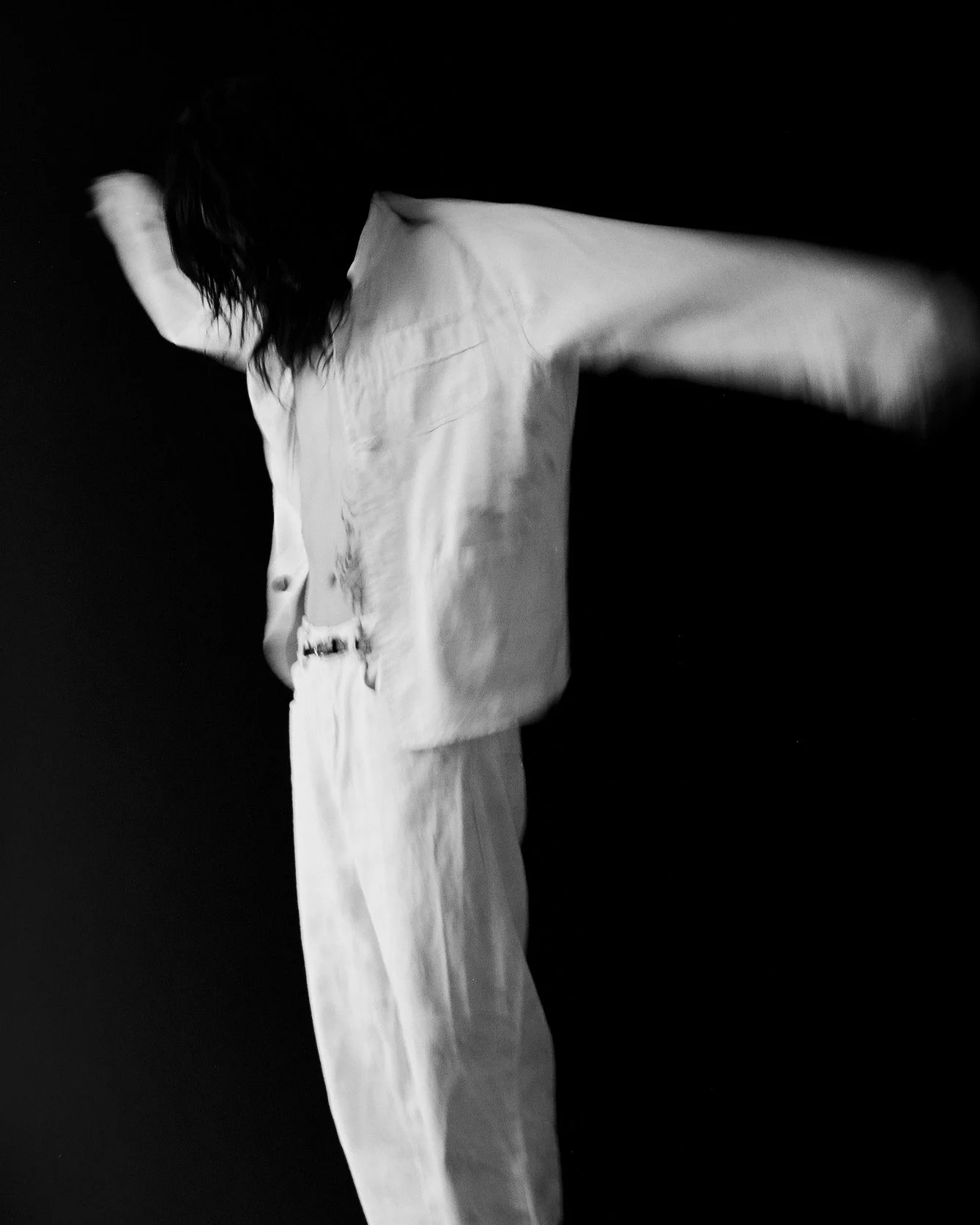 Black and white photo of a person with long dark hair wearing light-colored clothing, their head turned away from the camera, with one arm extended outward against a dark background.