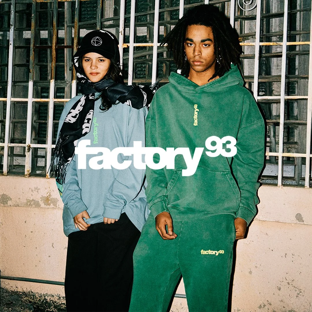 Two young individuals standing in front of a barred window, wearing casual streetwear with 'factory93' branding. The person on the left has long dark hair, wearing a cap, scarf, and oversized light blue jacket. The person on the right has dreadlocks, wearing a green hoodie and matching green pants.