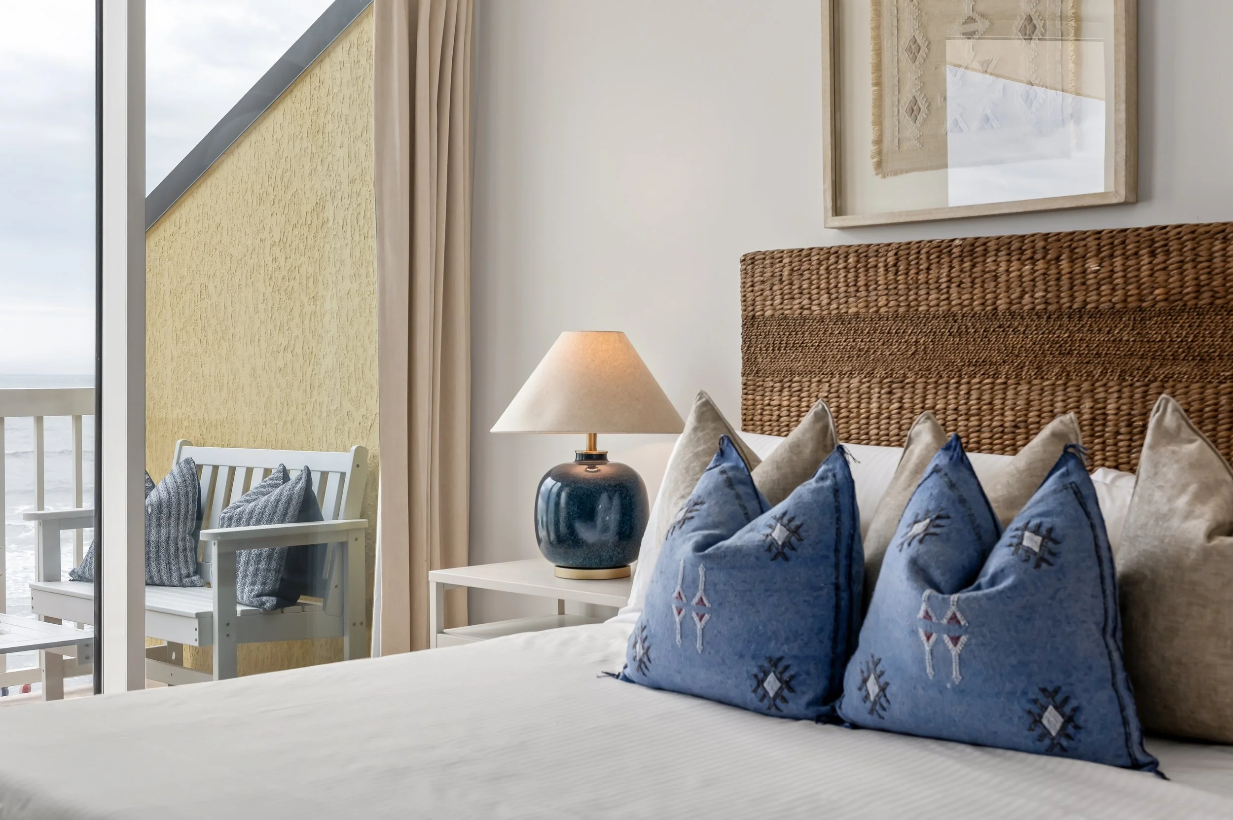 A cozy hotel bedroom with a bed, a wicker headboard, multiple blue embroidered throw pillows, a white nightstand with a dark blue lamp, beige curtains, a framed textile wall decor, and a balcony with white chairs and sea view.