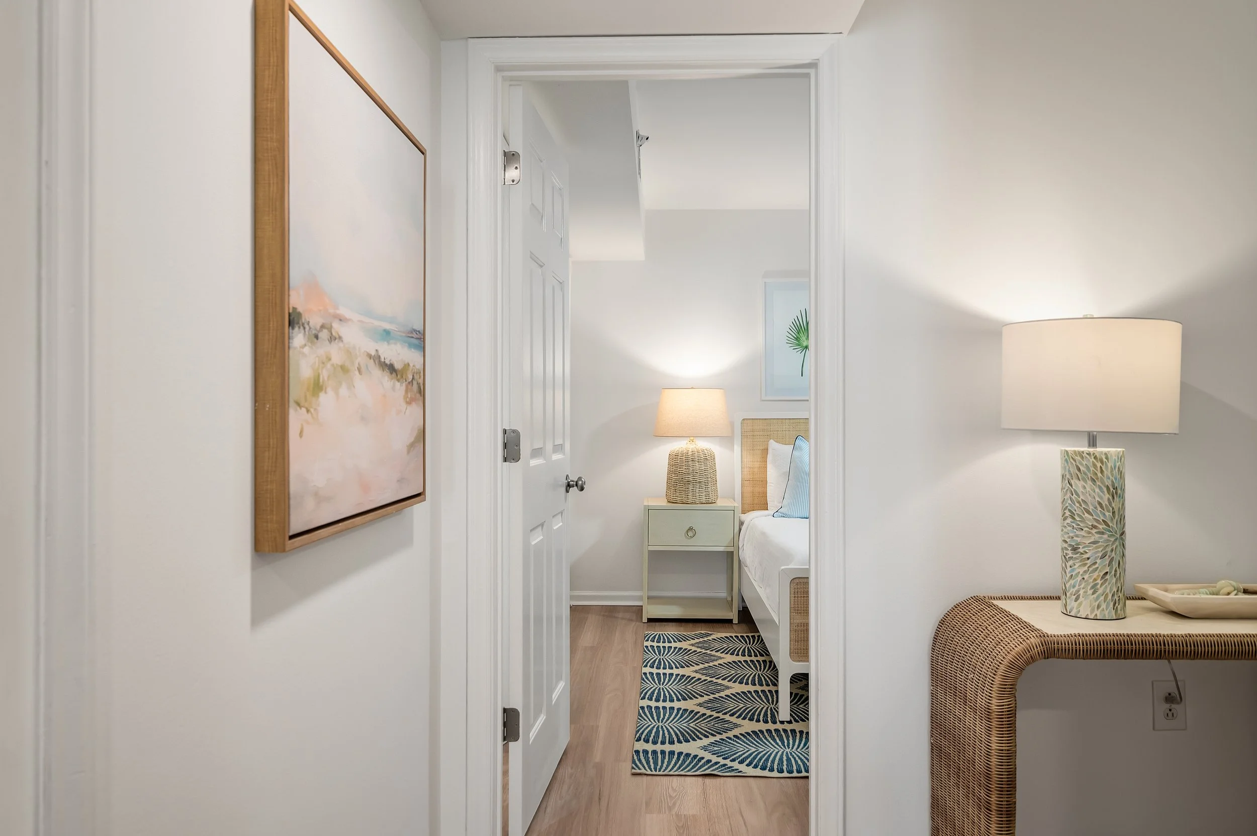 View into a bedroom showing a bedside table with a lamp, a bed with light colored bedding, a window with a plant, and a decorative rug on wood floor.
