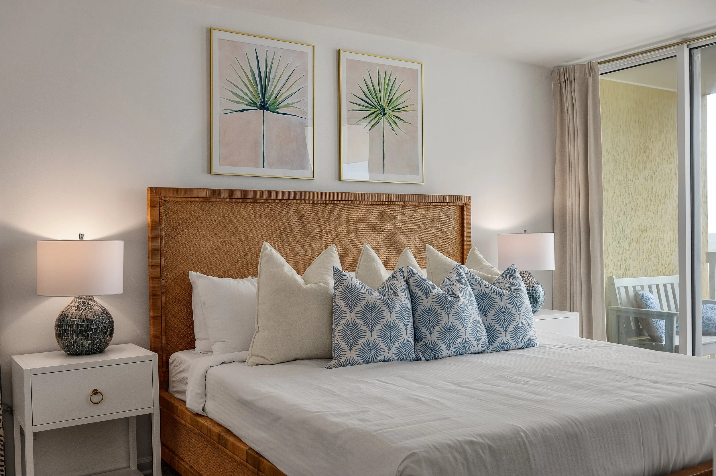 A neatly made bed with a rattan headboard in a modern bedroom. The bed has white linens, white pillows, and several decorative pillows with blue and white leaf patterns. There are white nightstands on each side of the bed, each with a textured gray and white lamp. Above the bed, there are two framed botanical prints of green leaves. To the right, there is a sliding glass door with beige curtains leading to a balcony with outdoor seating.