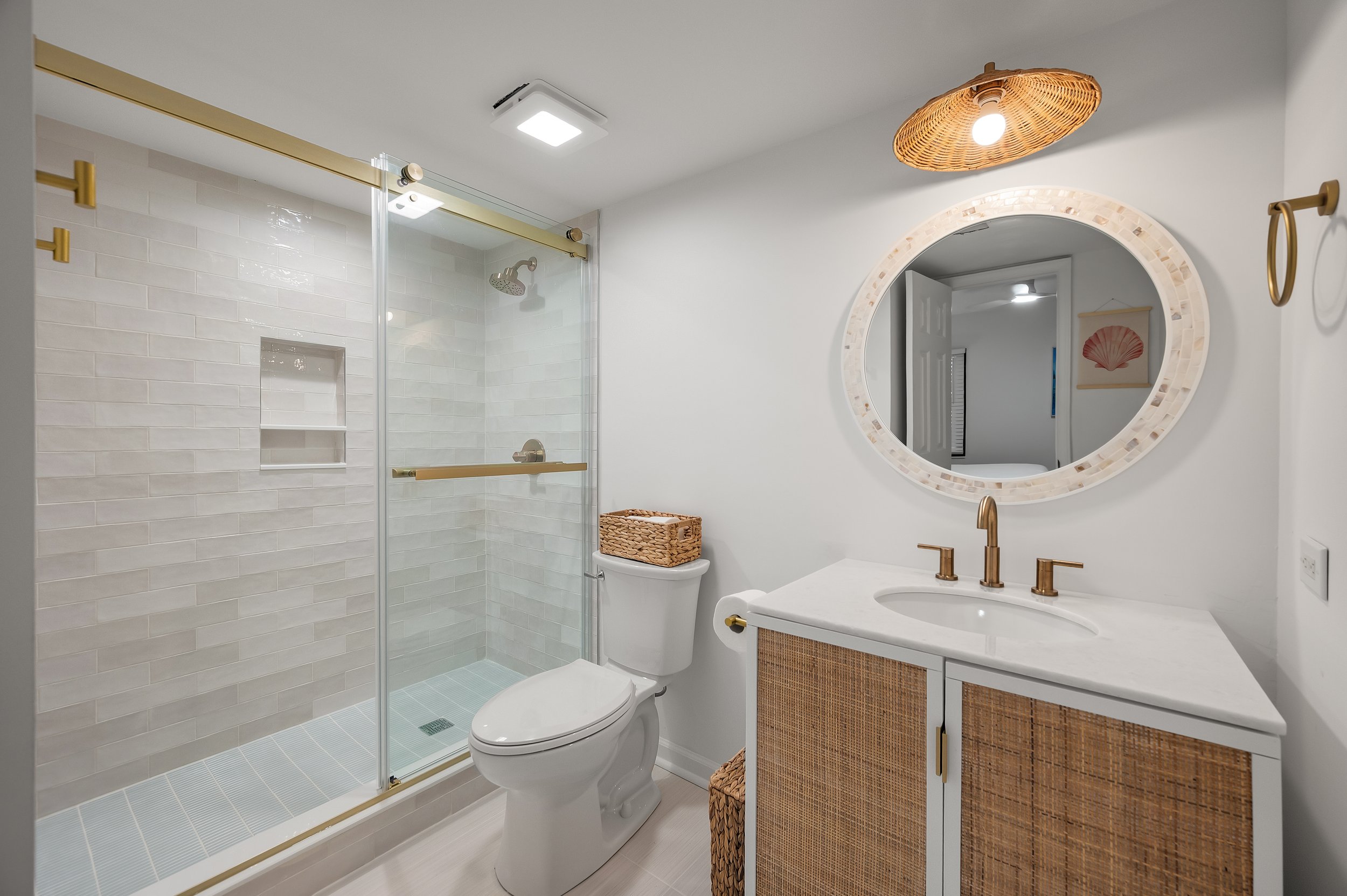 Modern bathroom with walk-in shower, white tiles, and a white toilet. A white vanity with wicker cabinet doors, a round mirror, and a wicker light fixture are above the sink. Decor includes a small wicker basket and a framed art piece of a seashell.