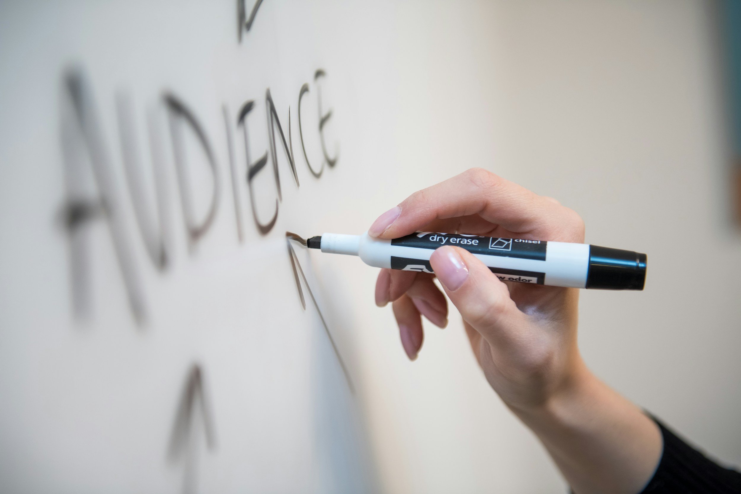 Person writing the word 'ADDIENCE' on a whiteboard with a black and white dry erase marker.