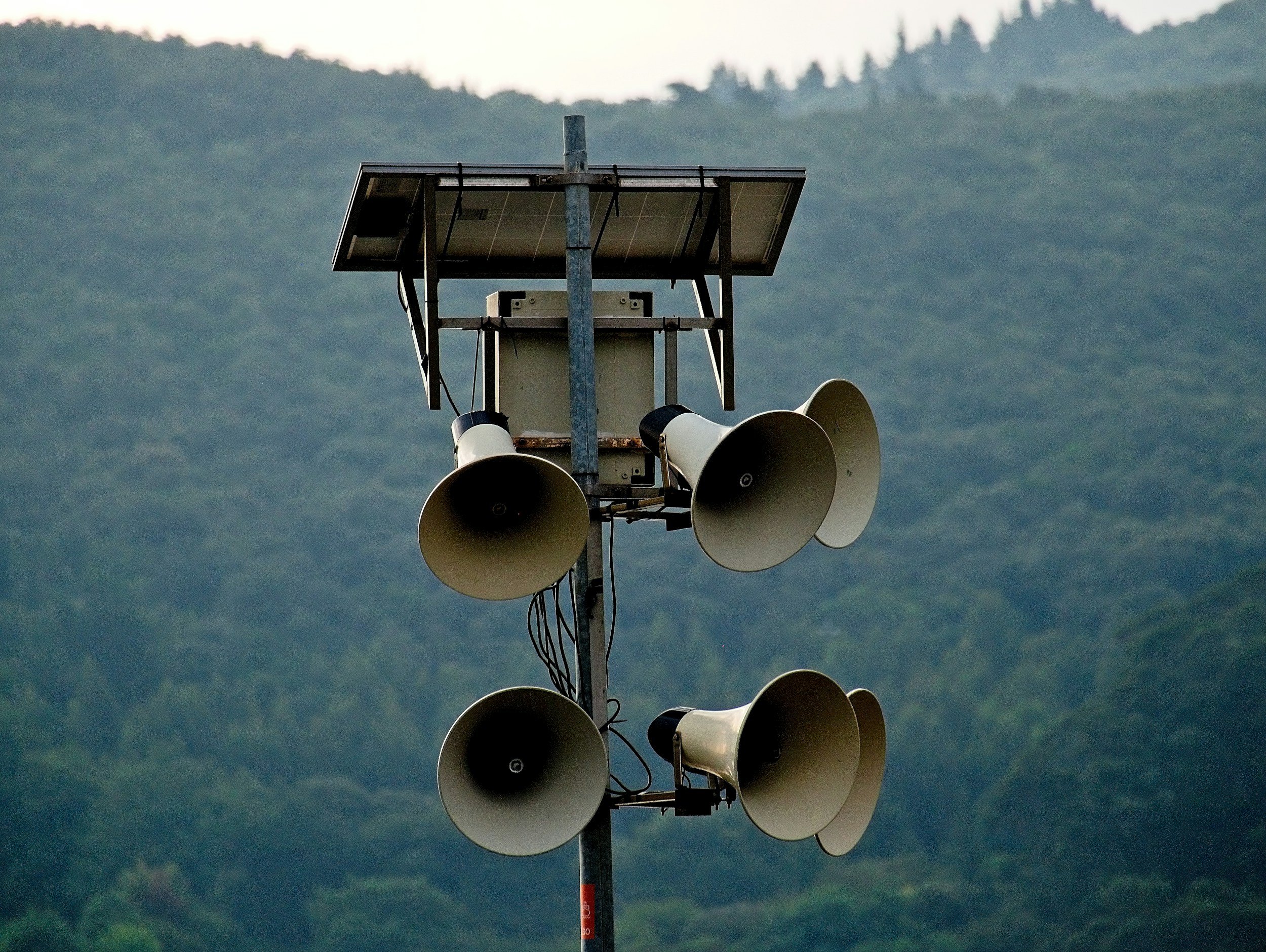 A tall radio tower with six large white megaphones and a solar panel on top, set against a backdrop of green, hilly mountains.