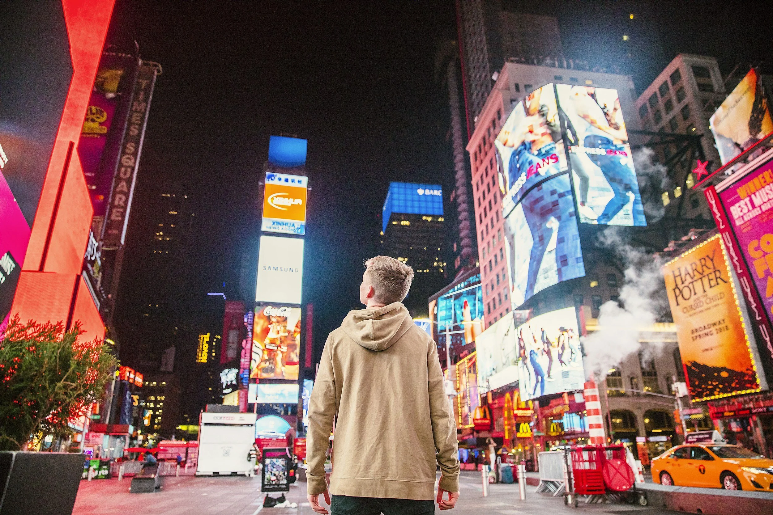 A person standing in Times Square, New York City at night, surrounded by brightly lit digital billboards and advertisements, with a street scene including taxis and storefronts.