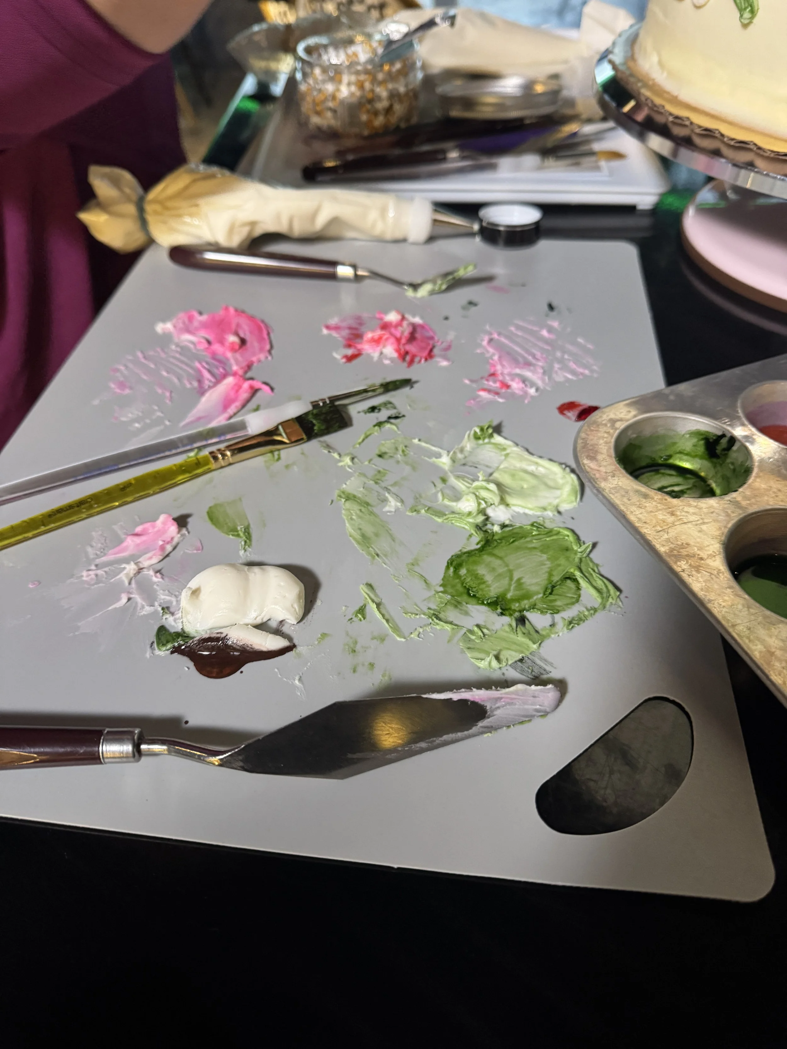 Paint palette with green, pink, and white paint, paintbrushes, and cake decorating tools on a work surface.