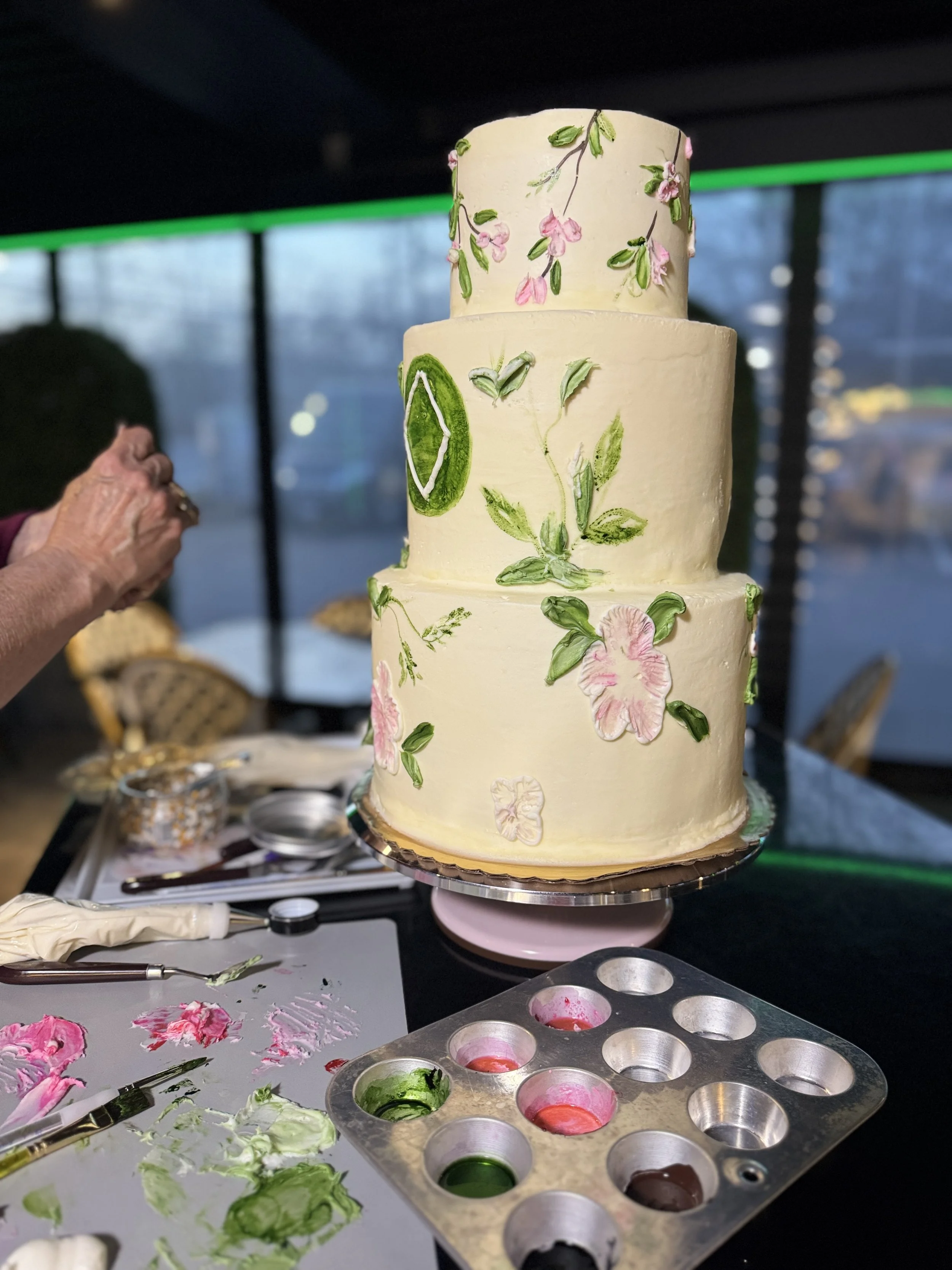 Three-tier white wedding cake decorated with pink flowers, green leaves, and a lime slice, displayed on a cake stand in a decorated room.