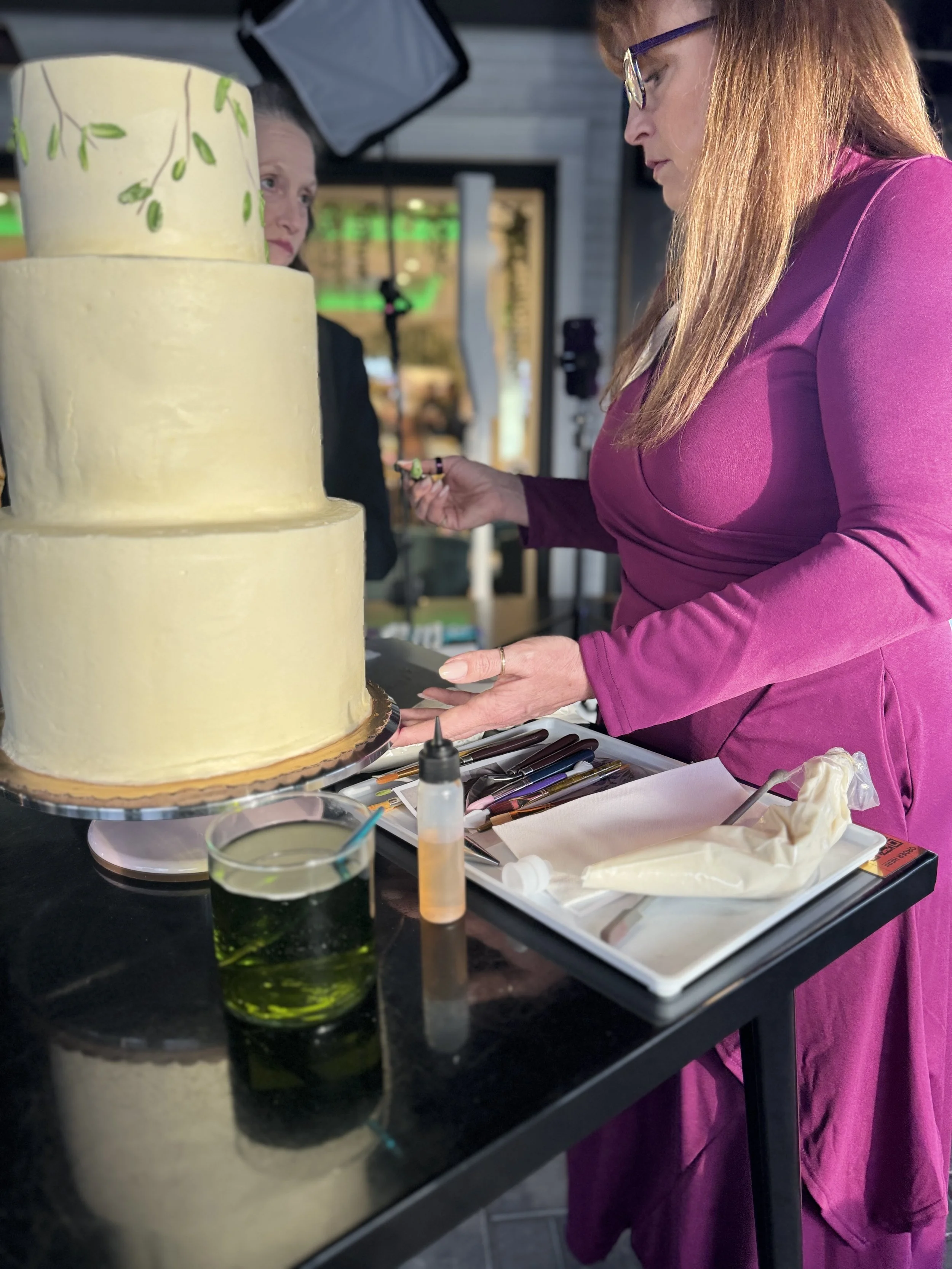 A woman in a magenta dress decorating a tiered cake at a cake decorating station with various piping tools and ingredients, in an indoor setting.