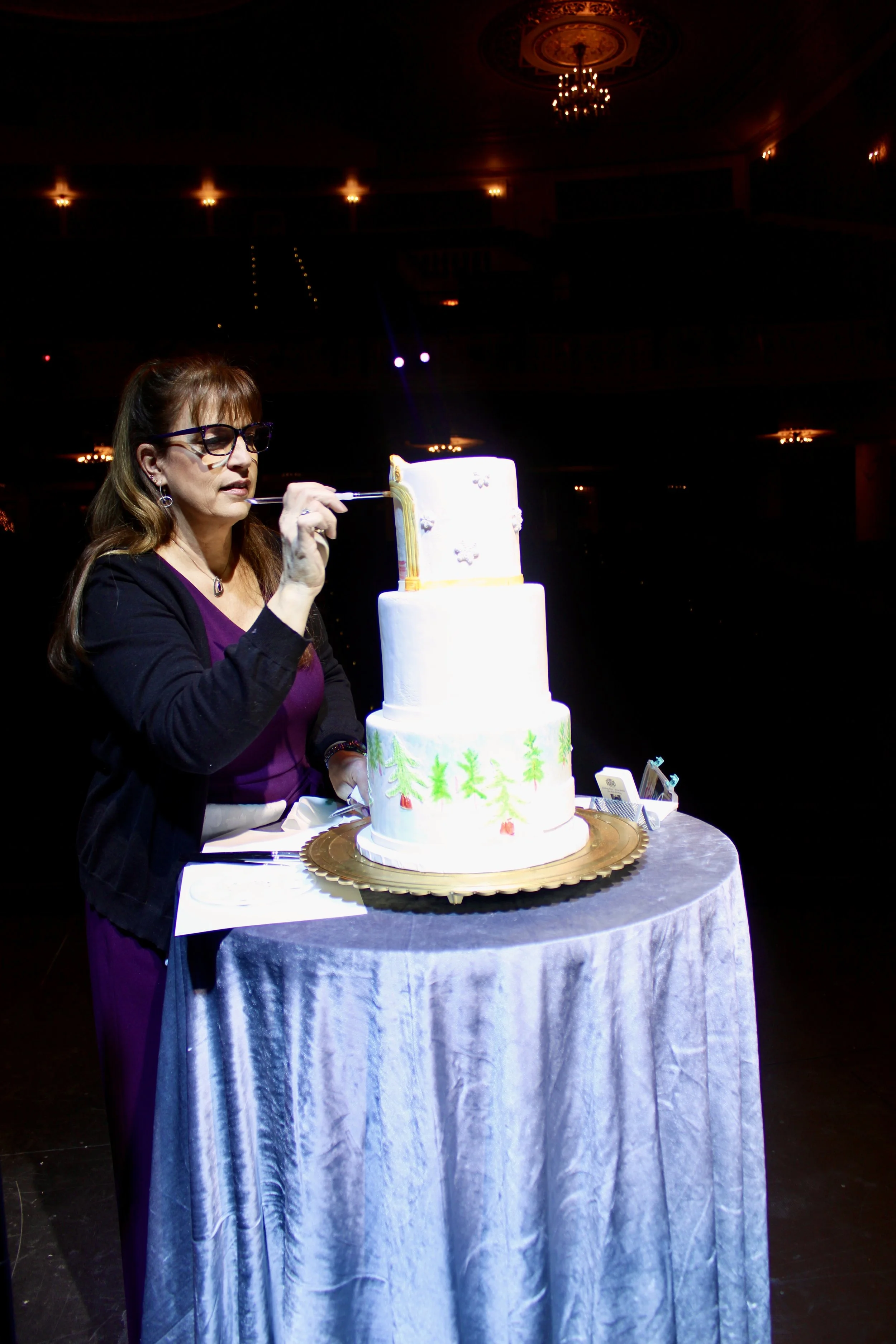 Woman with glasses decorating a three-tiered white wedding cake with a small piping tool at a silver-covered table in a dimly lit ballroom.