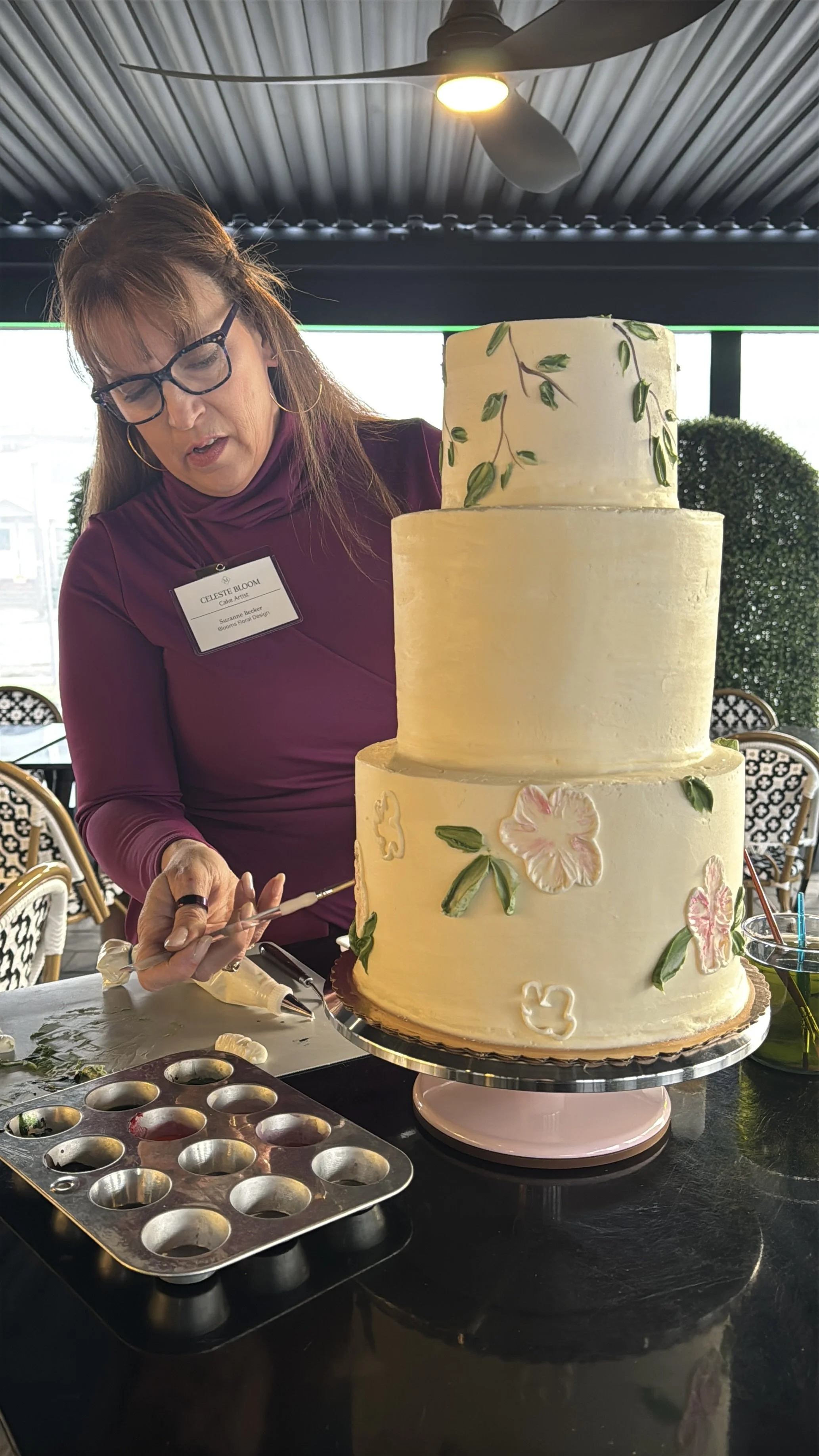 A woman with glasses in a purple top decorating a large, three-tiered white cake with floral designs at a table, with a cake decorating tools and a muffin tin nearby.