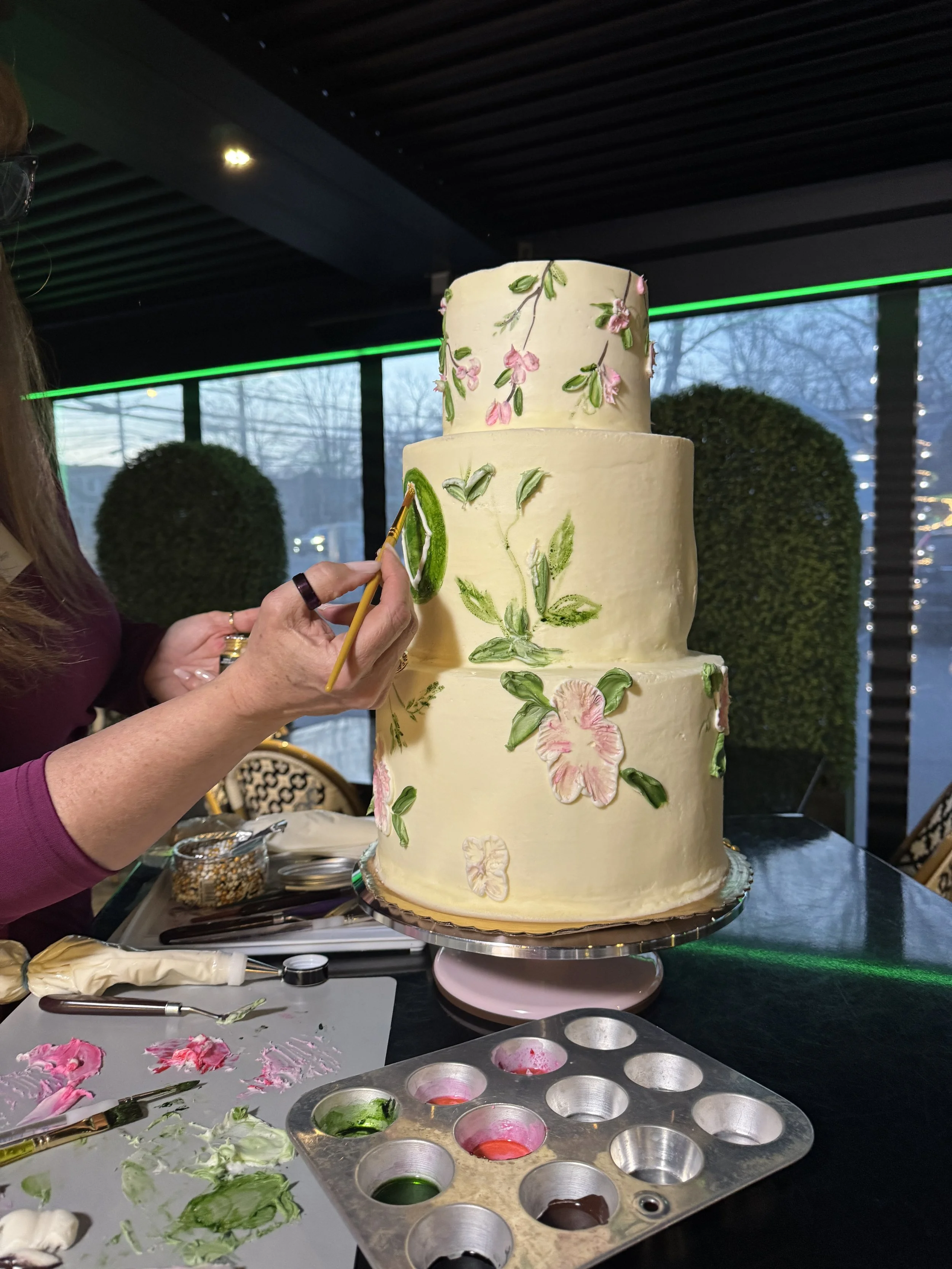 A person is decorating a multi-tiered cream-colored wedding cake with pink flowers and green leaves using a small paintbrush in a bakery or event space with large windows and natural light.