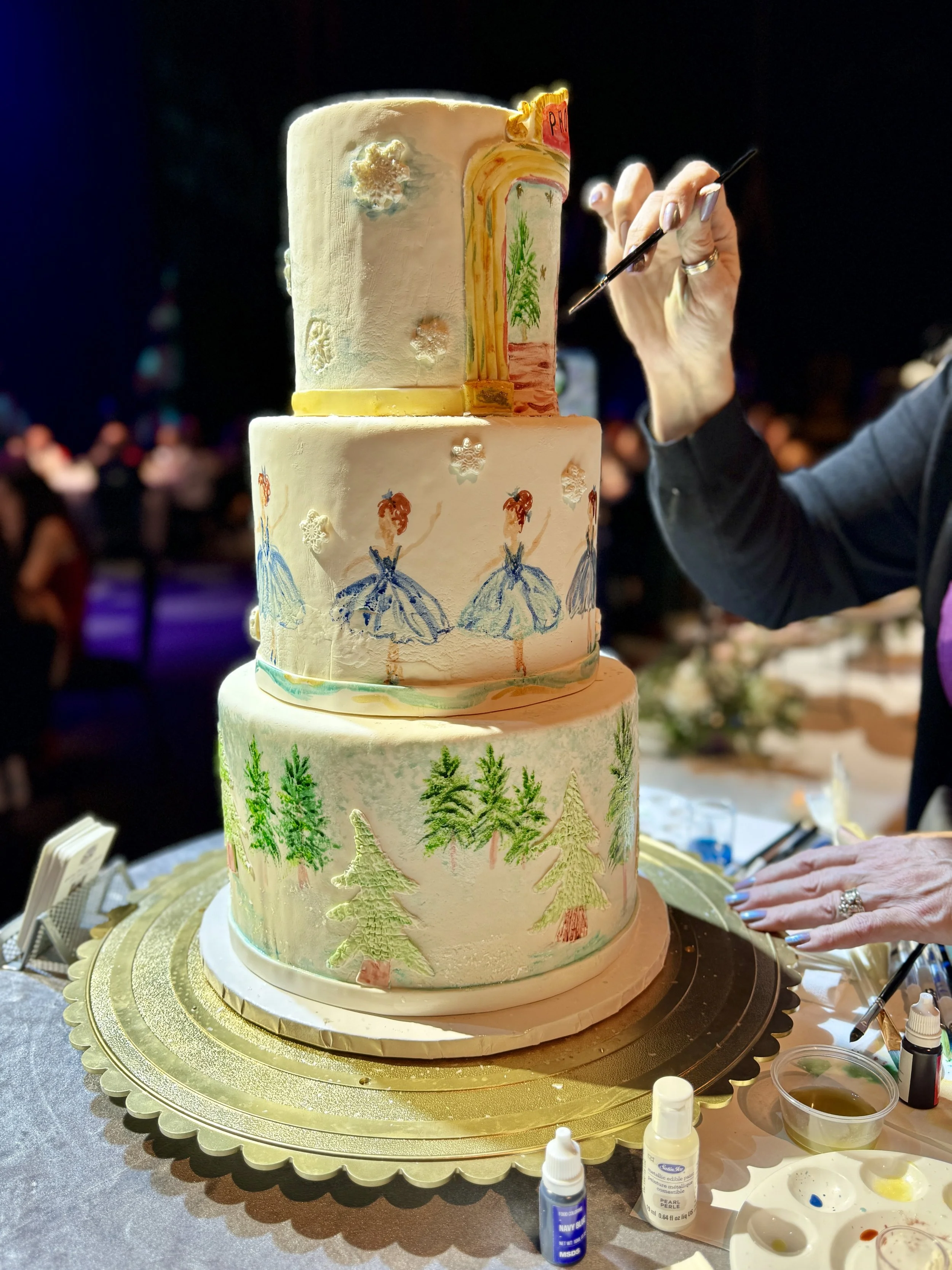 Three-tiered wedding cake decorated with painted trees, forest scenes, and ballet dancers, being decorated with a paintbrush by a person in dark clothing at a celebration event.