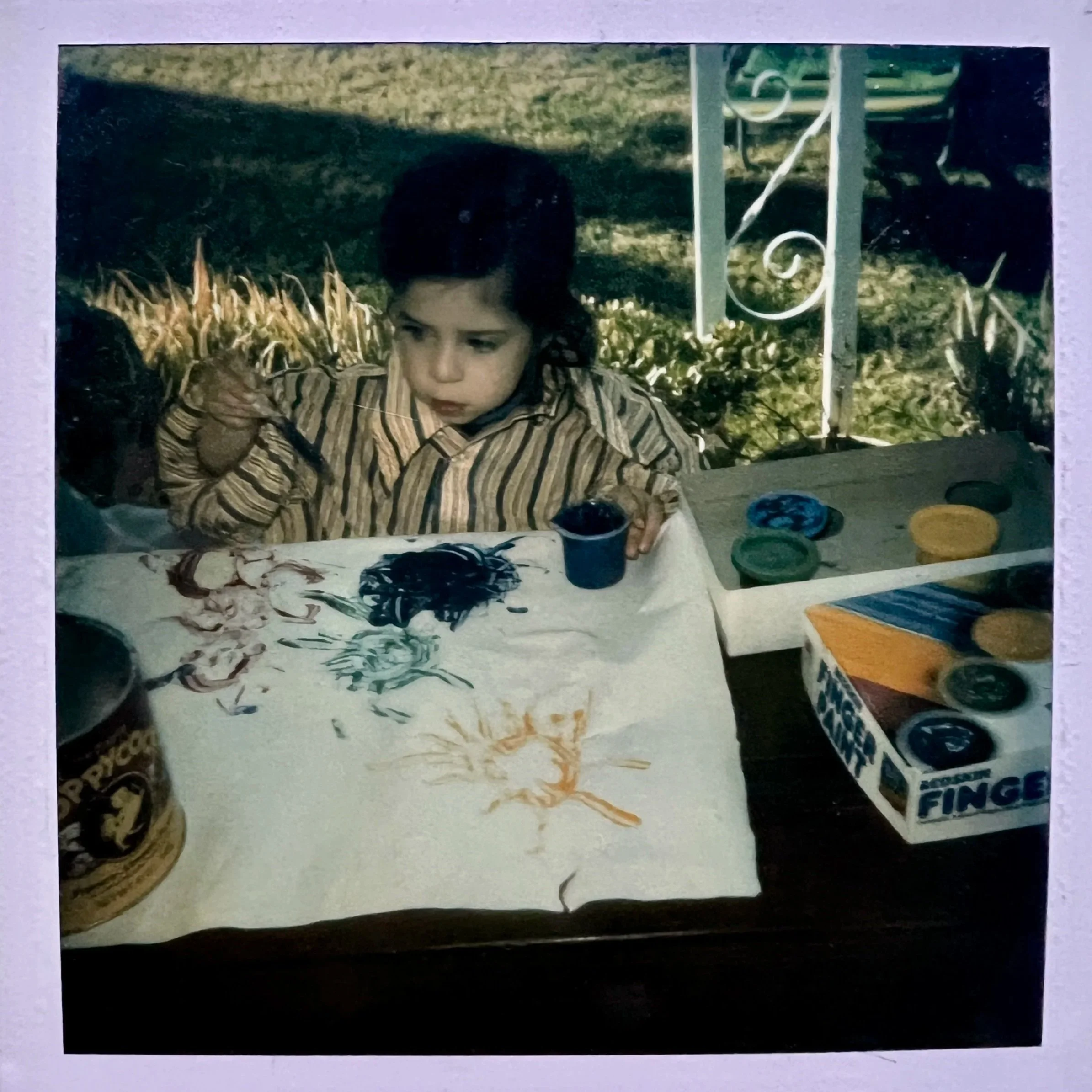 A young child with dark hair and light skin coloring, holding and looking at a paintbrush, wearing a vertically striped beige and brown shirt, is sitting at a table with paint supplies. Behind her is a lawn both and white metal vertical structure.