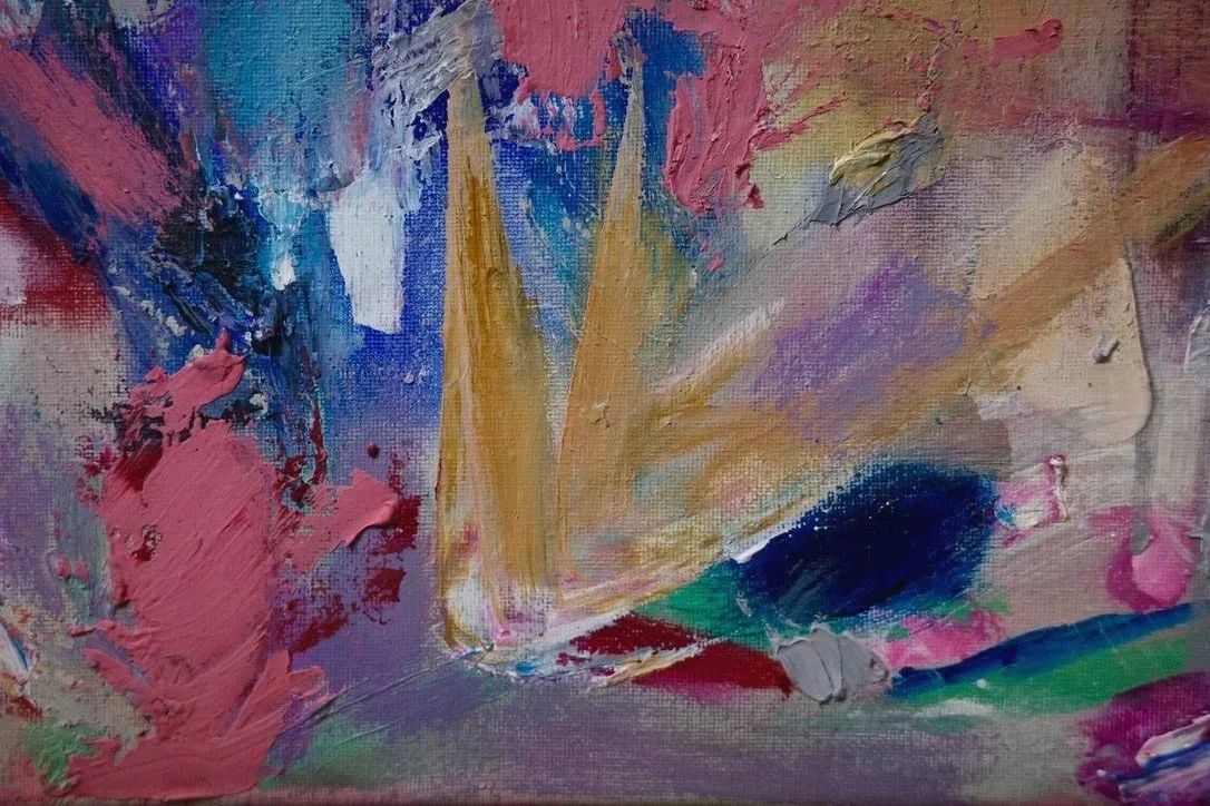 A close up of an abstract painting revealing thick, organic shapes and shades of pink, reds, blues, and yellows. Three muted yellow and white triangle-like shapes are at the center of the composition.