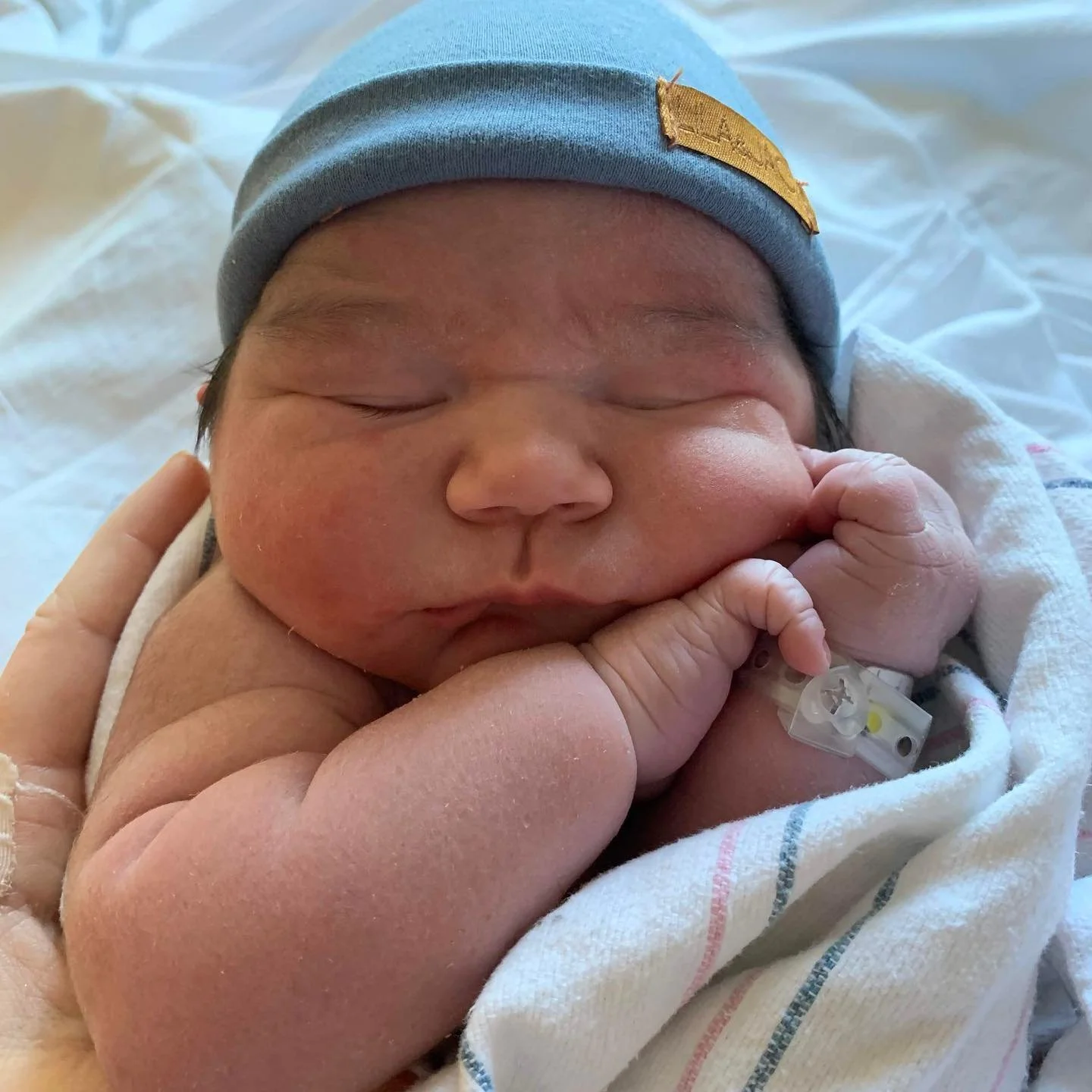 A newborn baby with a blue hat and hospital identification band, resting peacefully with its tiny hand near its face.