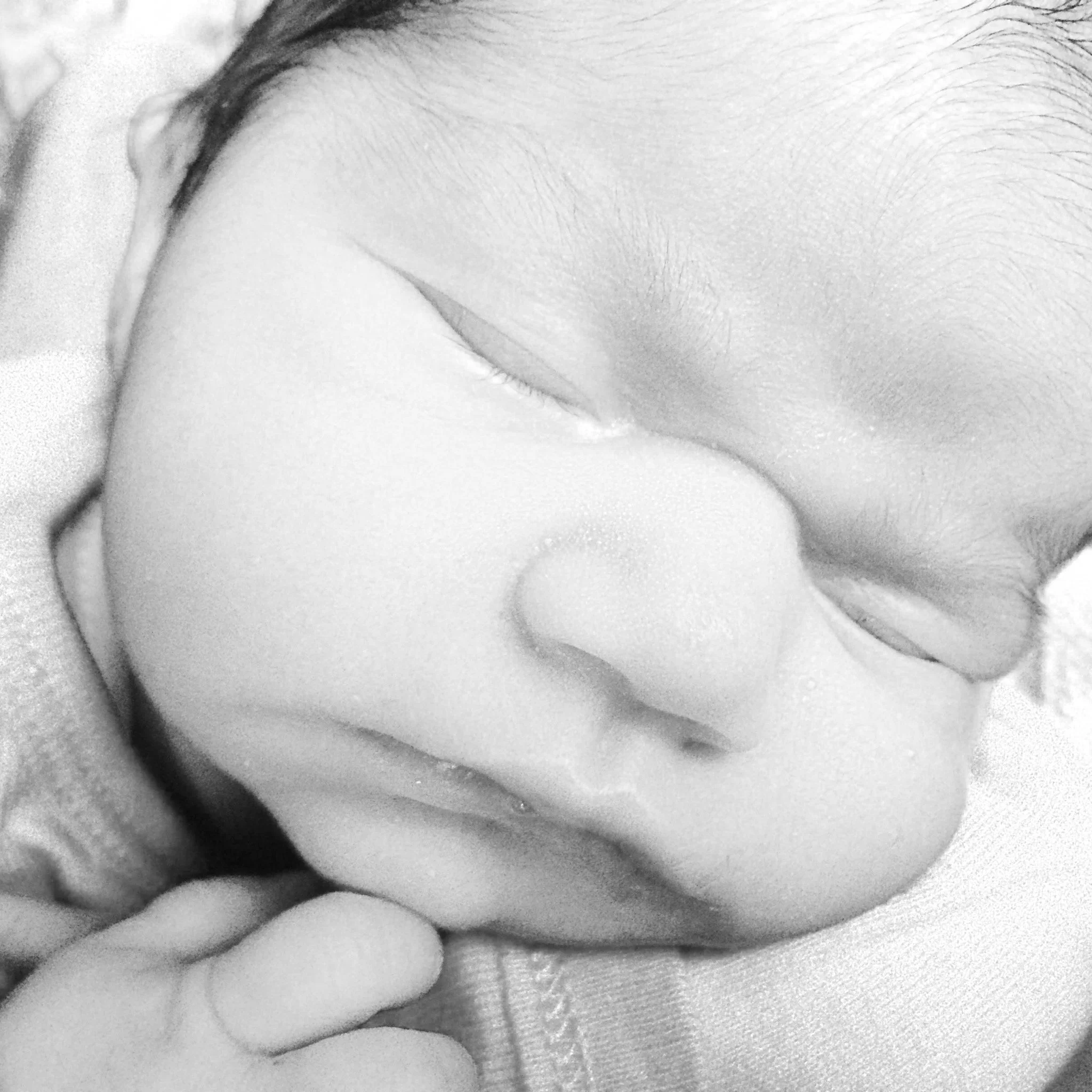 Close-up black and white photo of a sleeping baby with eyes closed, gentle expression, and soft skin, resting their cheek on their hand.