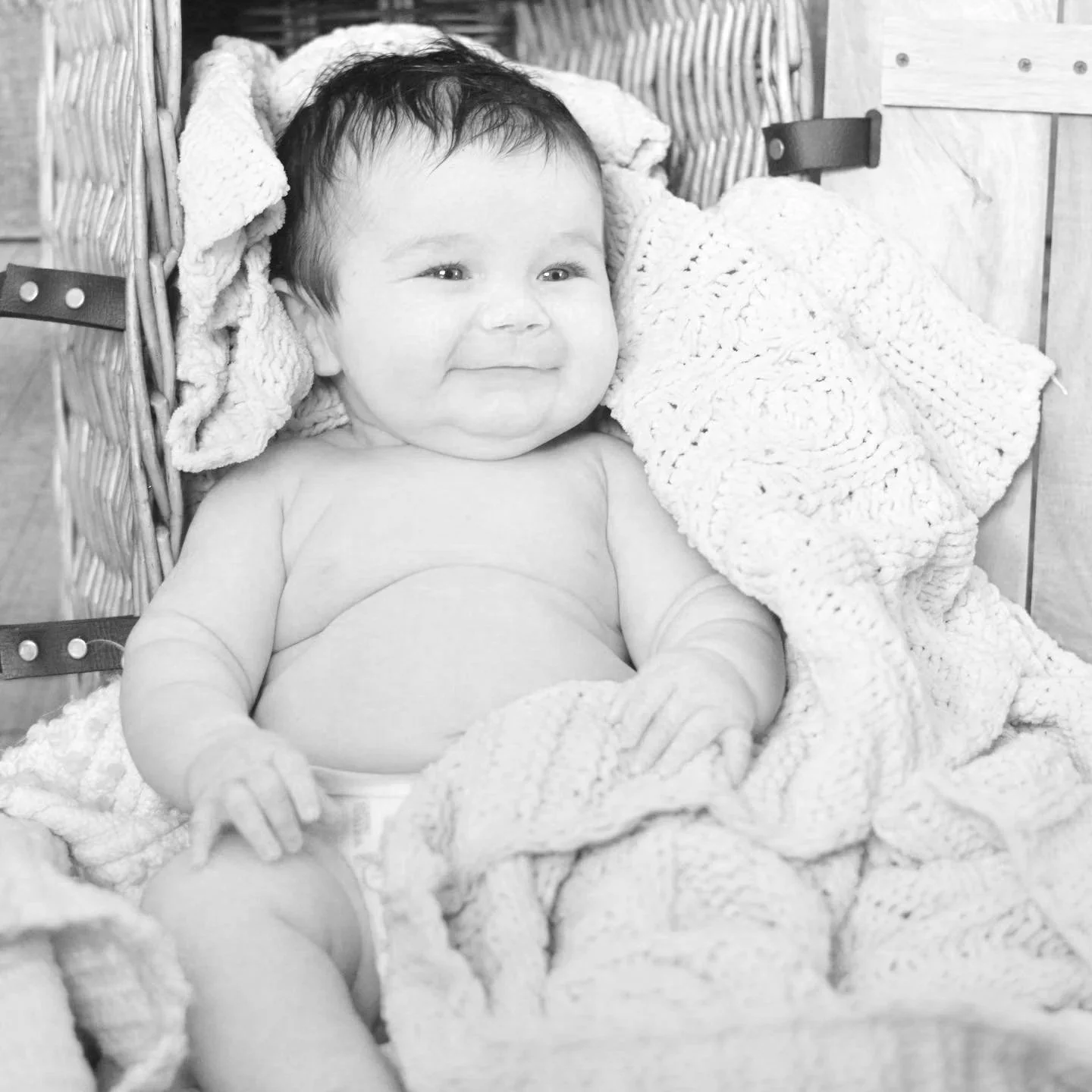 A smiling baby sitting in a wicker basket with a knitted blanket.