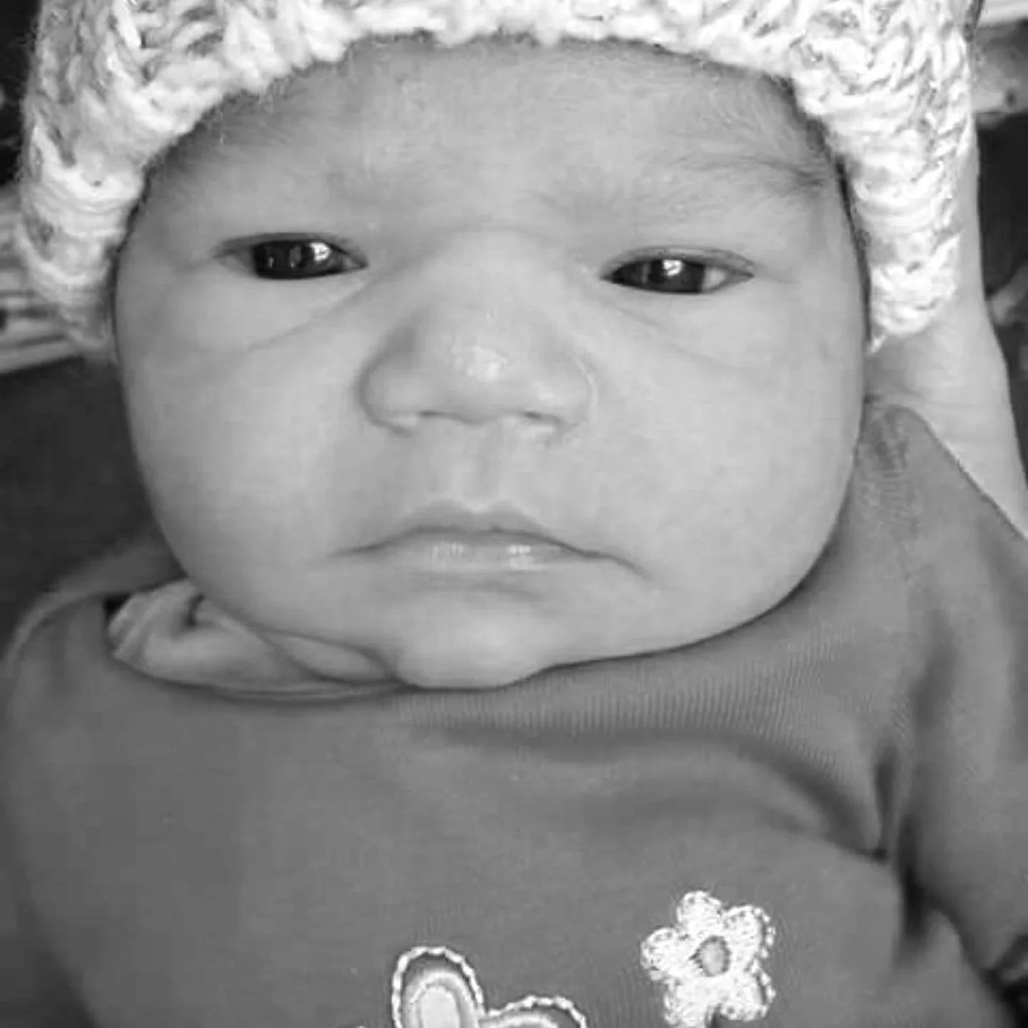A close-up black and white photo of a baby's face wearing a knit hat and a shirt with flower and heart designs.