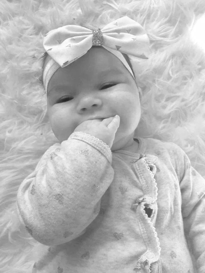 A baby lying on a fluffy surface, wearing a headband with a large bow and a long-sleeve shirt, smiling and touching her mouth with her hand.